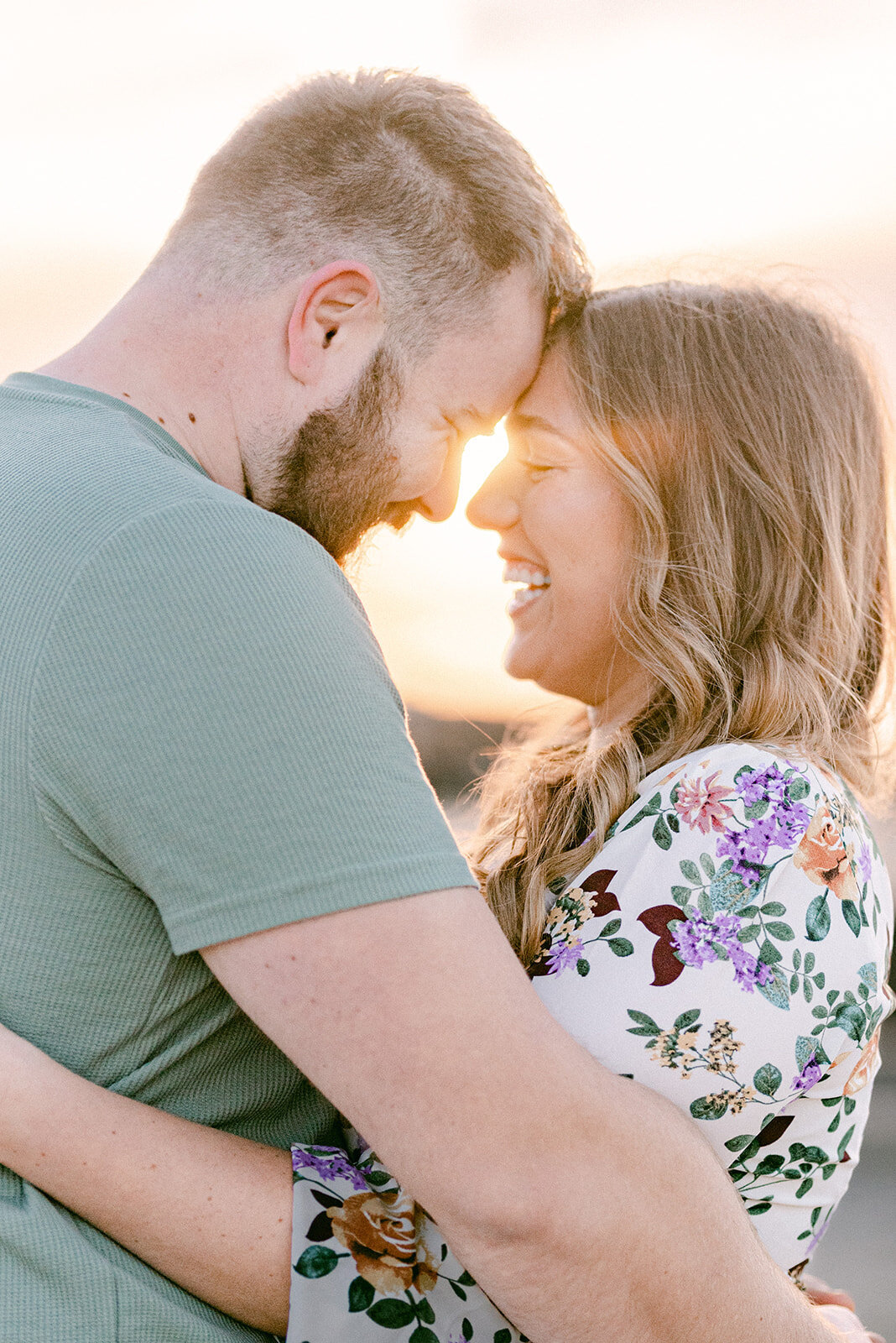 Sunrise engagement photoshoot at the beach, the couple have their foreheads together and are smiling at each other with the sun flaring between them