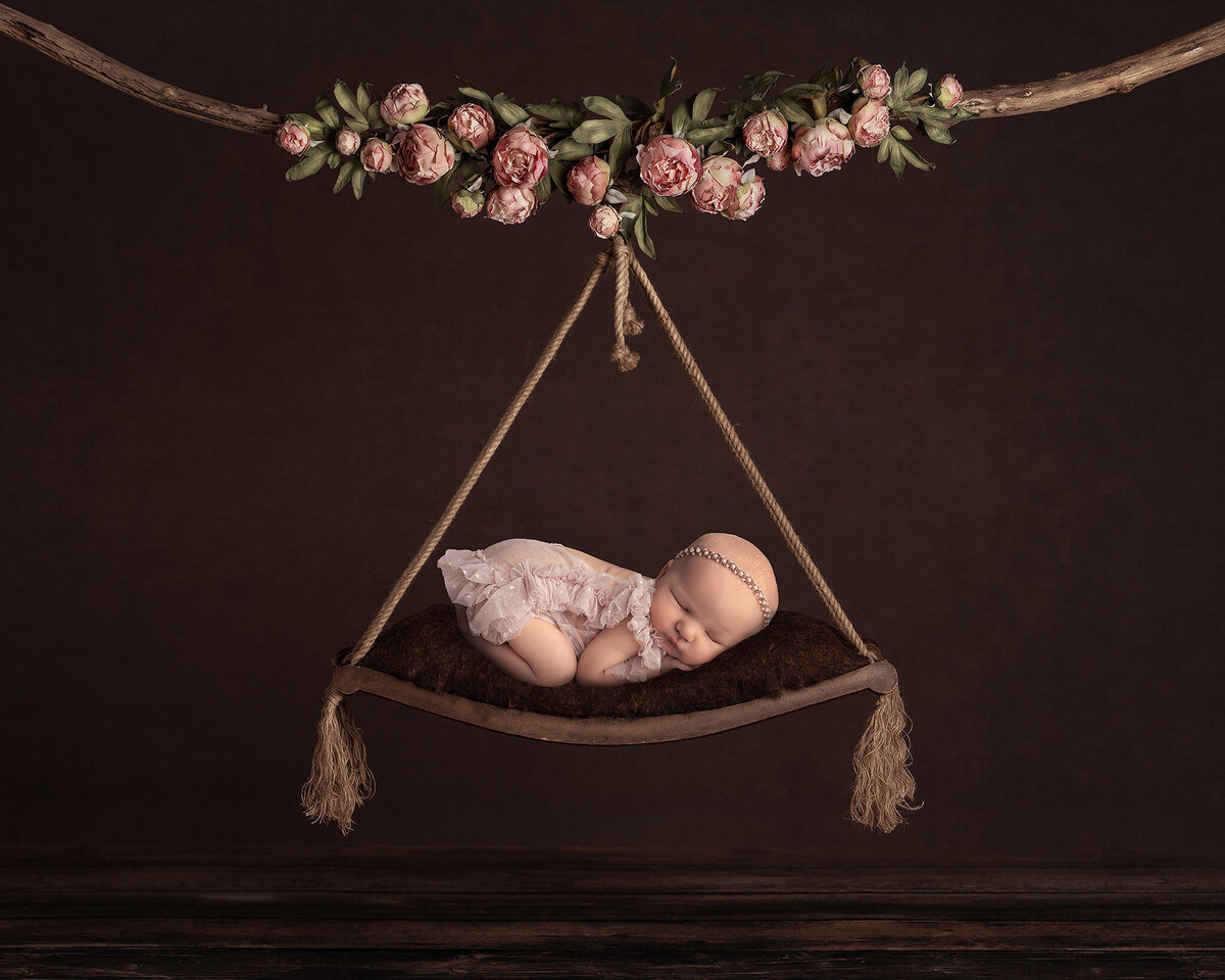 Newborn baby girl in a pink ruffled romper posed on a suspended floral swing, beautifully styled by a Dallas baby photographer specializing in newborn pictures and artistic newborn photography.