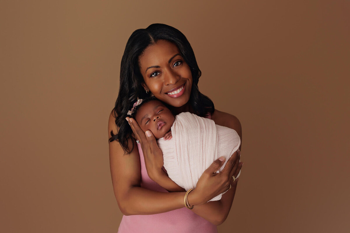 A smiling mother in a pink dress gently holding her newborn baby swaddled in white against a brown background.