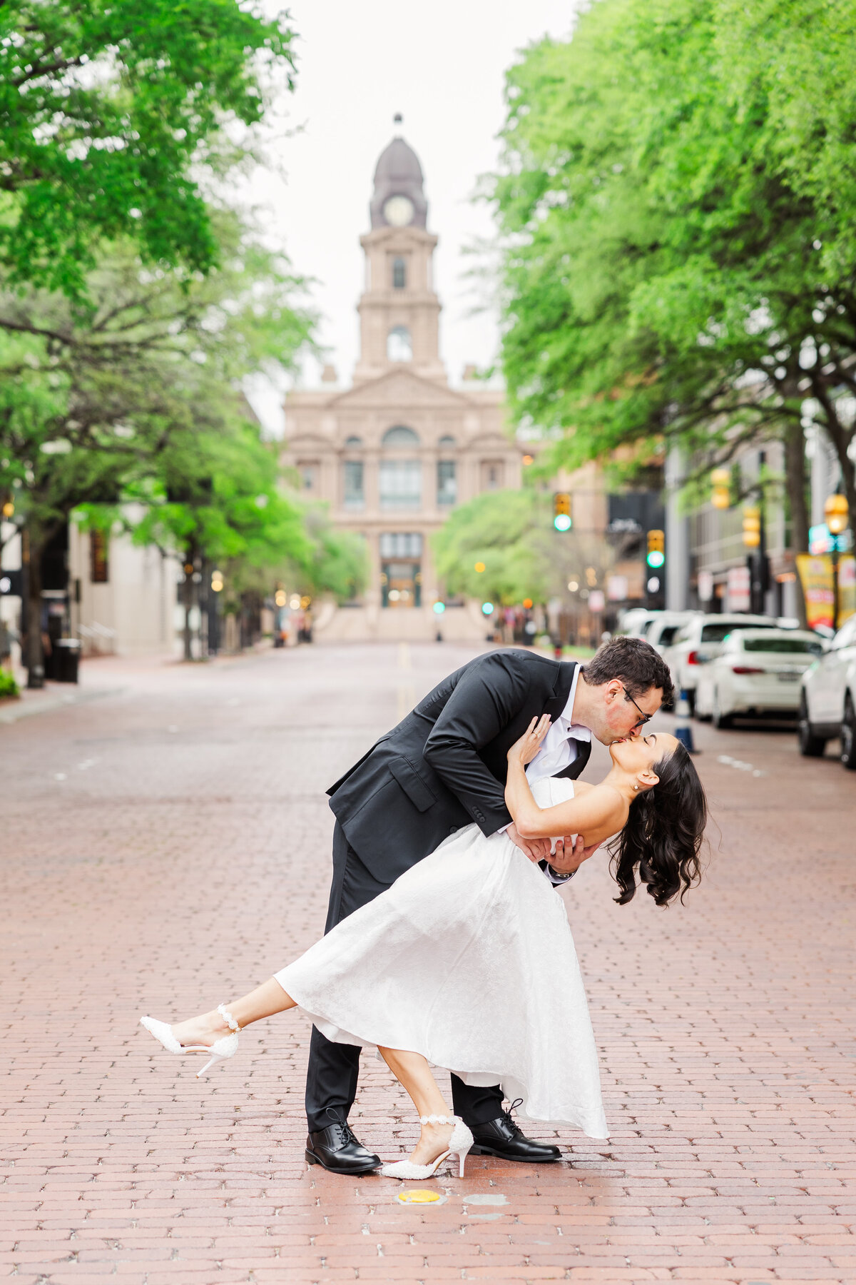 Chic engagement photo of a couple sharing a romantic dip-and-kiss moment on a brick-paved street in downtown Fort Worth. The iconic courthouse in the background adds timeless Texas charm to this elegant and joyful scene.
