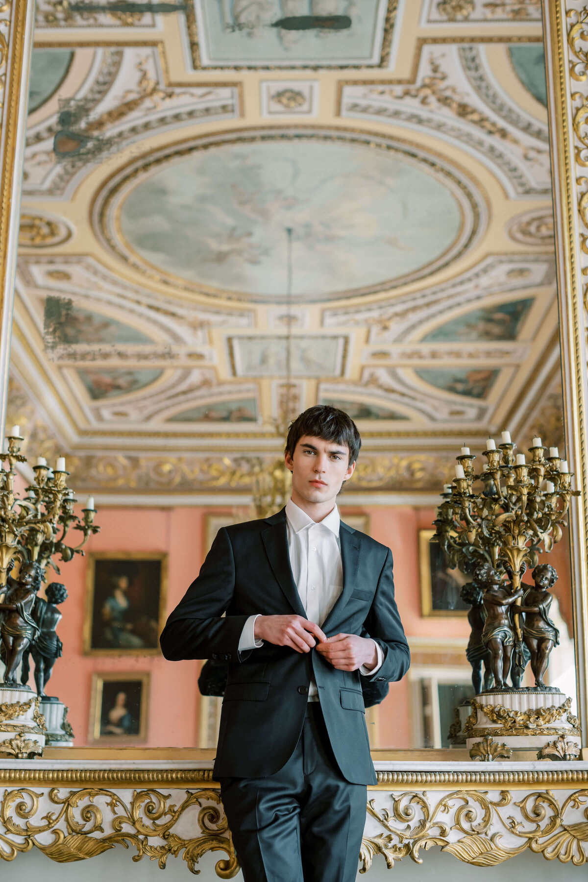 groom buttoning a black wedding suit in the versailles ballroom at avington park with a mirror behind him reflecting the elaborate ceiling painting
