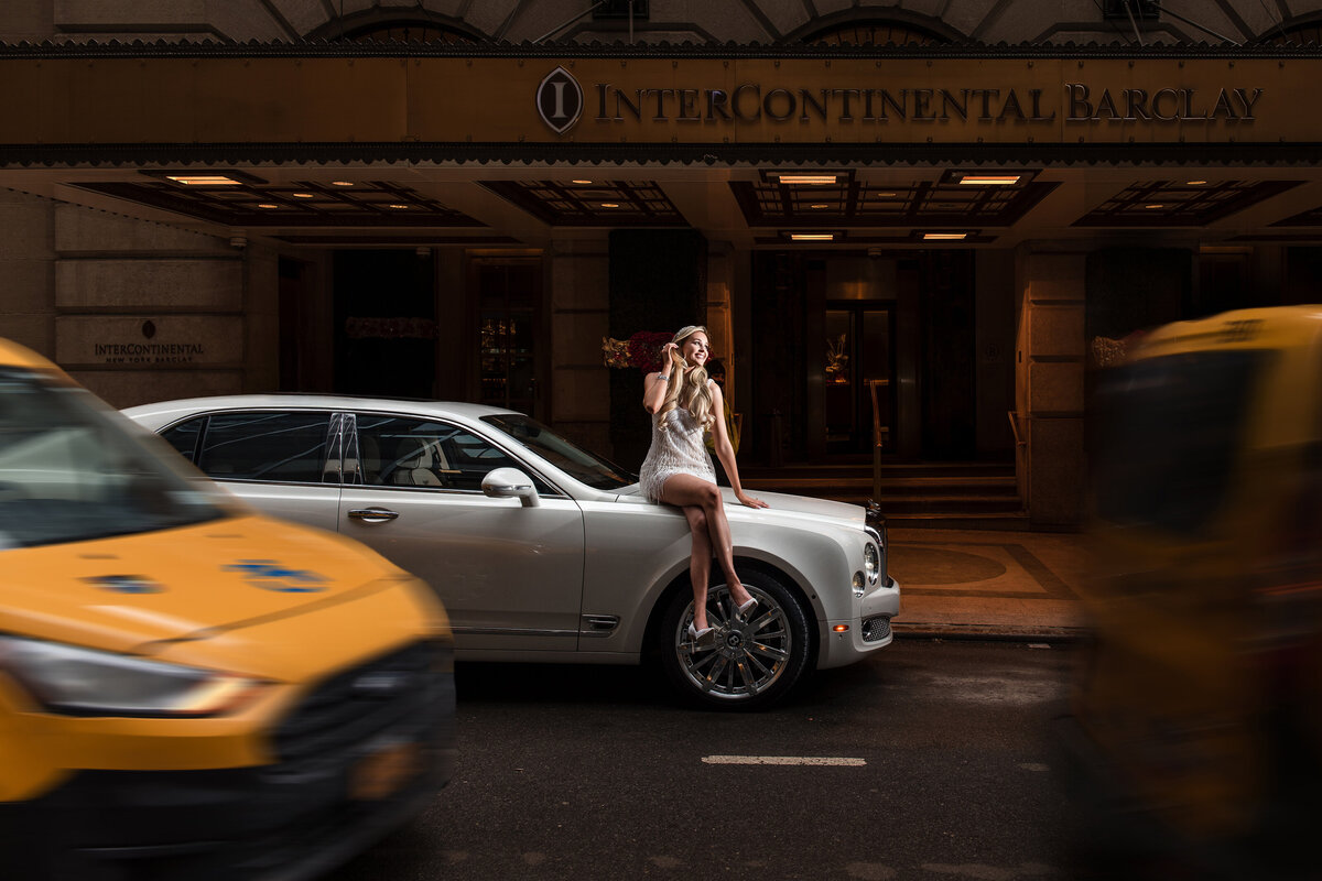 A stylish bride in a beaded dress poses on the hood of a white luxury car outside the InterContinental Barclay Hotel in Manhattan, with iconic NYC yellow cabs in motion on either side—capturing the energy of a luxury New York City wedding with editorial flair.