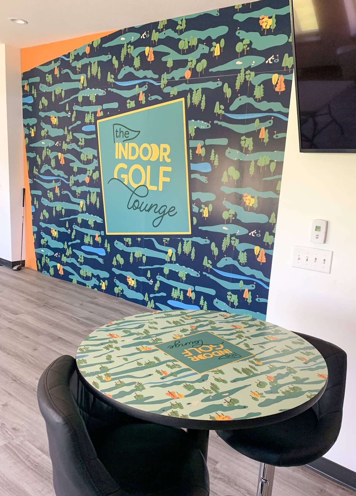A round table with The Indoor Golf Lounge brand pattern and logo printed on the top sits in front of a large wall featuring the same pattern and logo, but with a darker background color