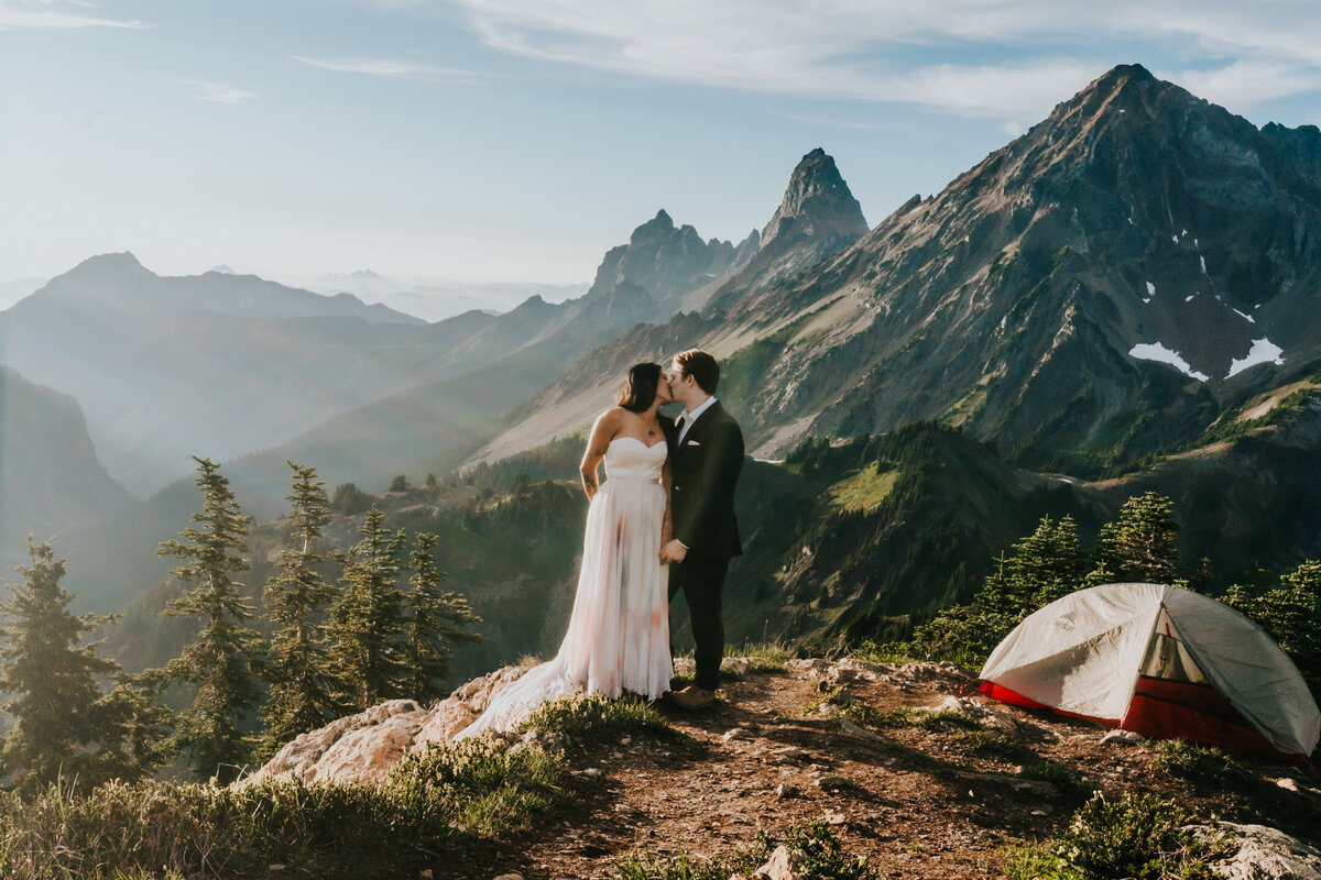 a couple in their wedding attire kiss with jagged mountains rising above em in the background. A backpacking tent can be seen in the frame as their adventure elopement photographers capture this moment from their elopement day