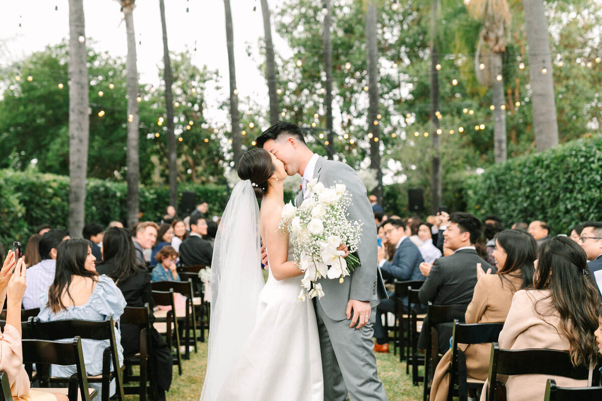 Bride in a white dress and groom in a gray suit share a first kiss outdoors. They're surrounded by applauding guests, string lights, and lush greenery.