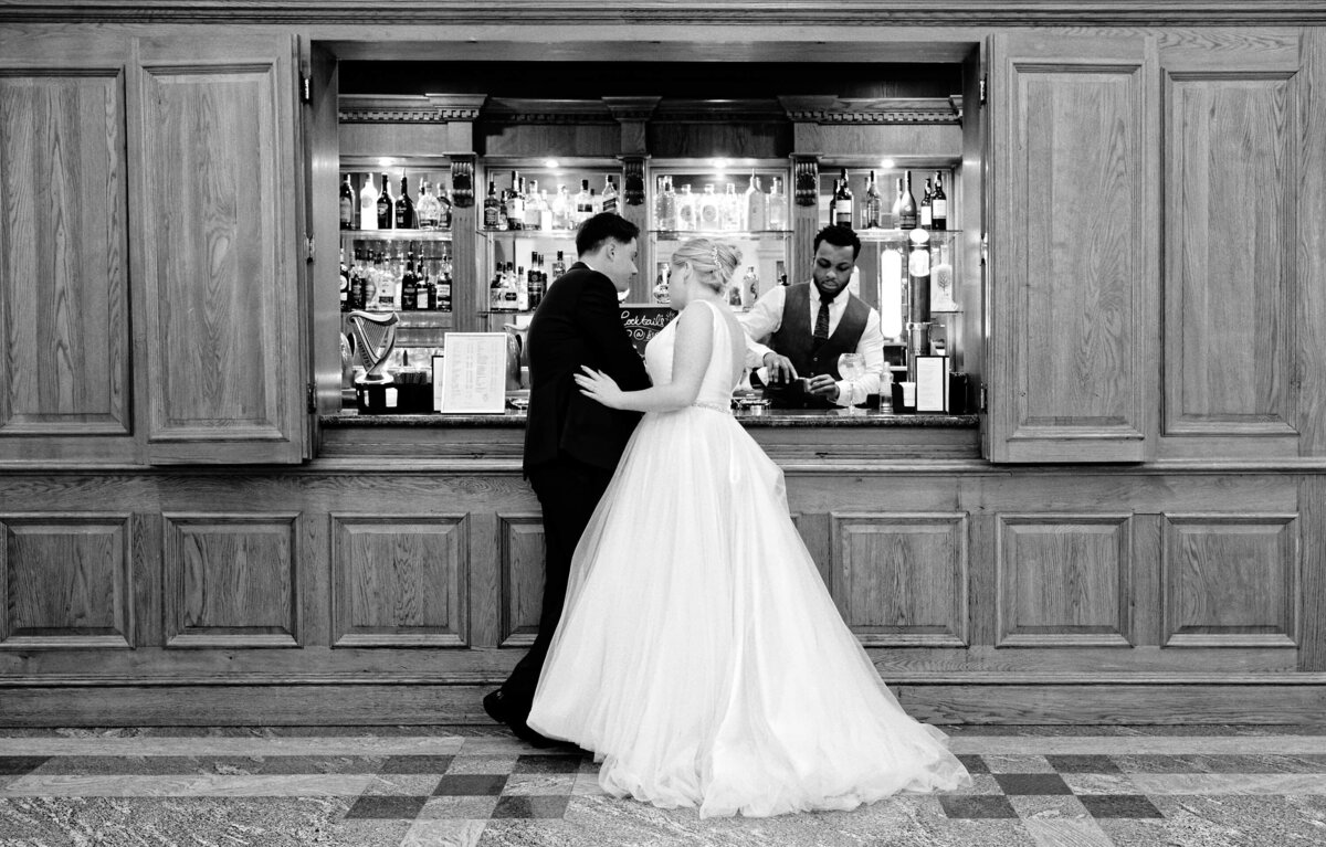 Black and white photo of a tender moment between a bride and groom while waiting for their drinks to be served by a barman at the bar in the South Lodge Hotel.