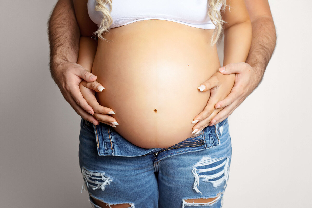 pregnant mother wearing jeans and a white sports bra with her and her husbands hands on her belly on a white backdrop at a maternity photo shoot in northern va