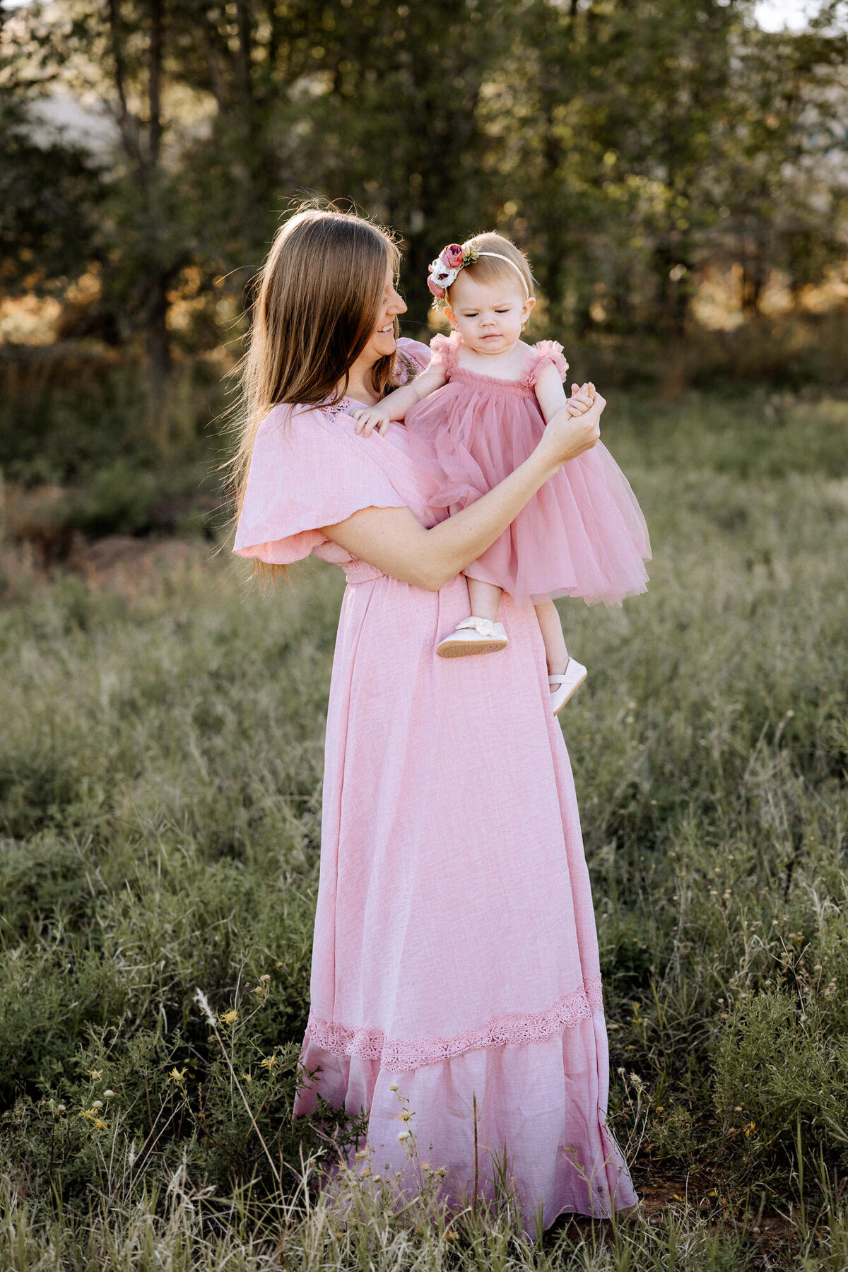 A mother in a flowing pink dress holds her young daughter in a matching pink tulle dress in an open field with lush greenery. The mother gazes lovingly at her child, who wears a floral headband and soft baby shoes. The golden sunlight filters through the trees, creating a warm and heartfelt moment captured in nature.