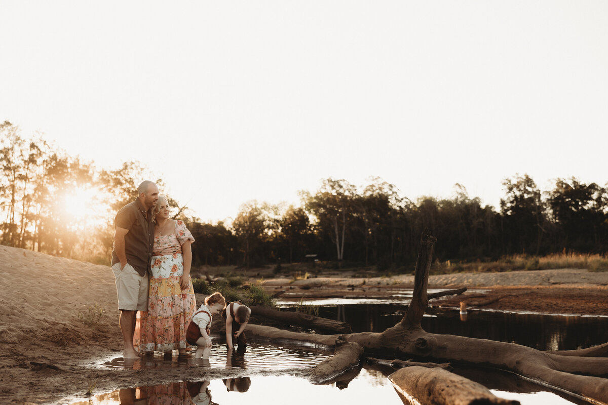 A family of four stands by a calm river at sunset. The parents watch as two children explore the water's edge. Fallen logs and trees are visible in the background—a perfect scene for South Coast family photography.