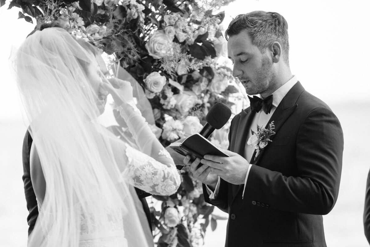 Ceremony on the beach with floral arch behind couple, bride is wiping away a tear as the groom holds the microphone reading vows