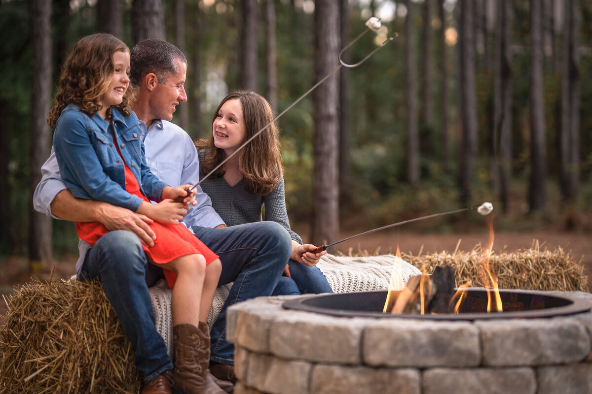 A dad in a blue shirt is sitting at a fire pit making smores with his daughters.