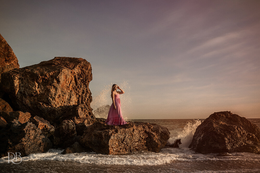 teen girl standing on a rock in a pink dress at the beach with the ocean behind her