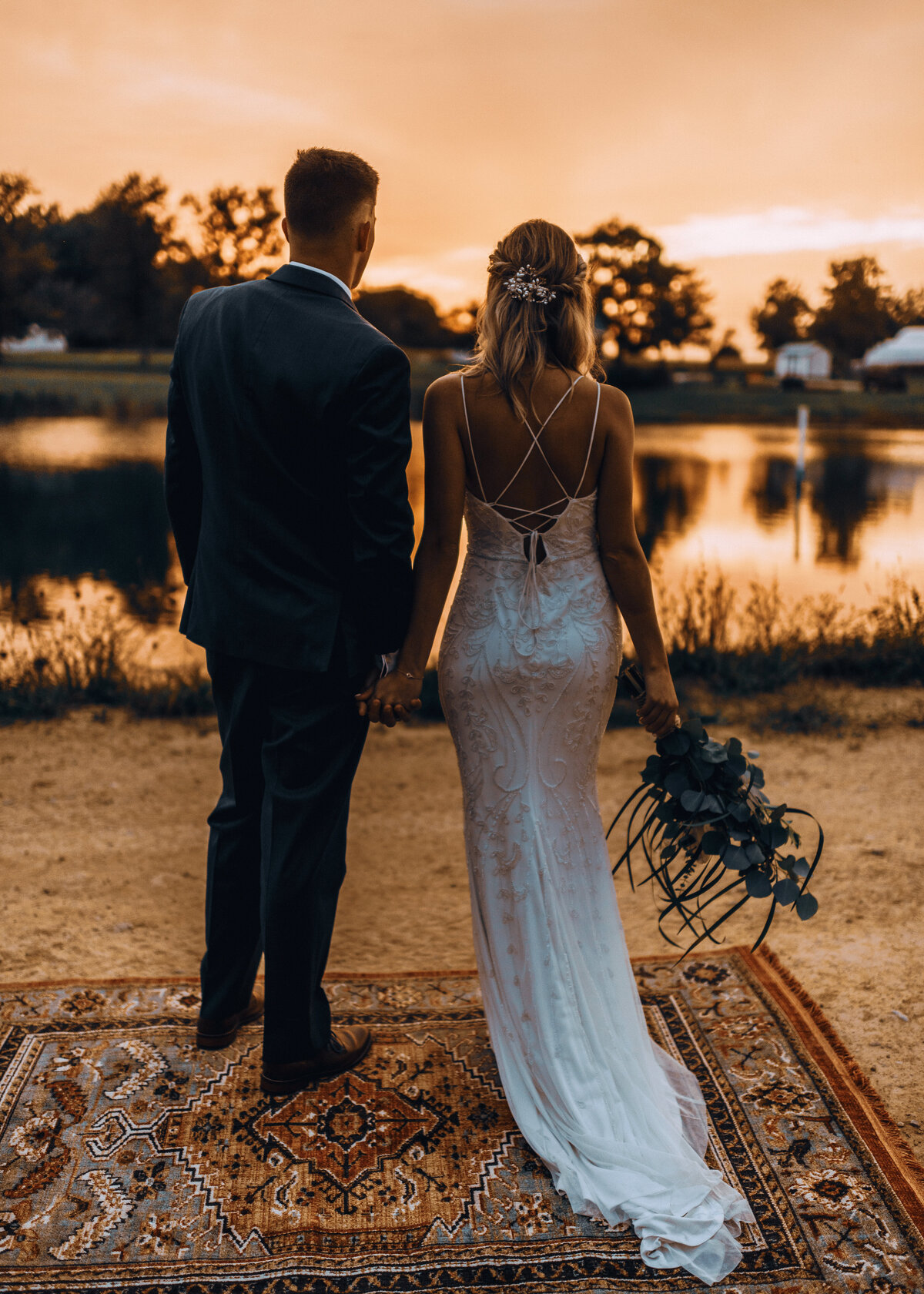 A boho wedding couple stand on a rug looking out over the lake. The bride has a backless dress.