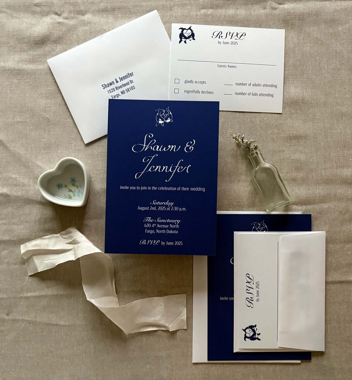 Custom calligraphy blue wedding suite with rsvp card and white envelope. Flatlay with silk ribbon, florals and heart dish