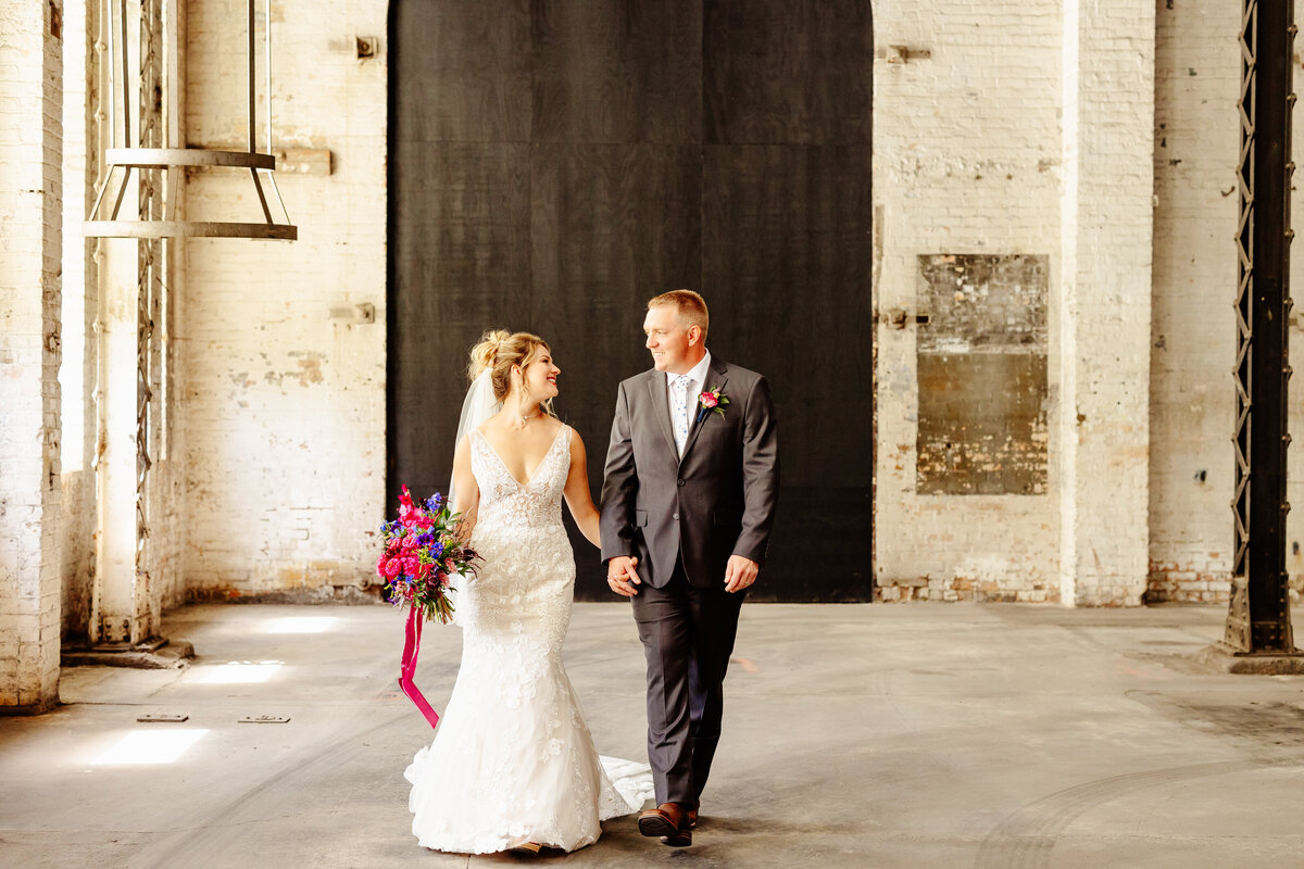 A bride in a white dress holds a colorful bouquet as she walks with her groom, who is dressed in a gray suit. Captured by a skilled Brainerd Lakes Area photographer, they smile at each other against the backdrop of an industrial setting with white brick walls and large black doors.