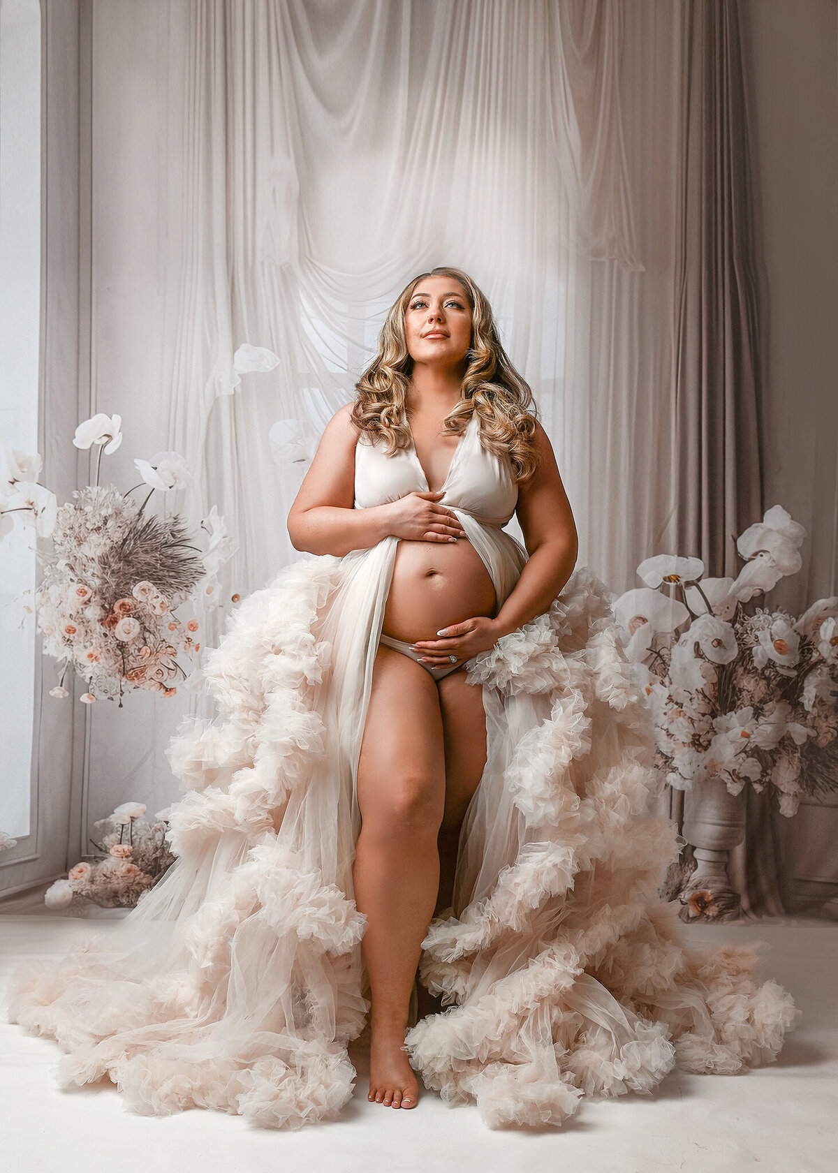 Exvpecting woman with long blond curled hair, wearing cream tulle gown, standing against a tan/cream windon and curtain background with floral arrangements in Scottsdale Maternity studio