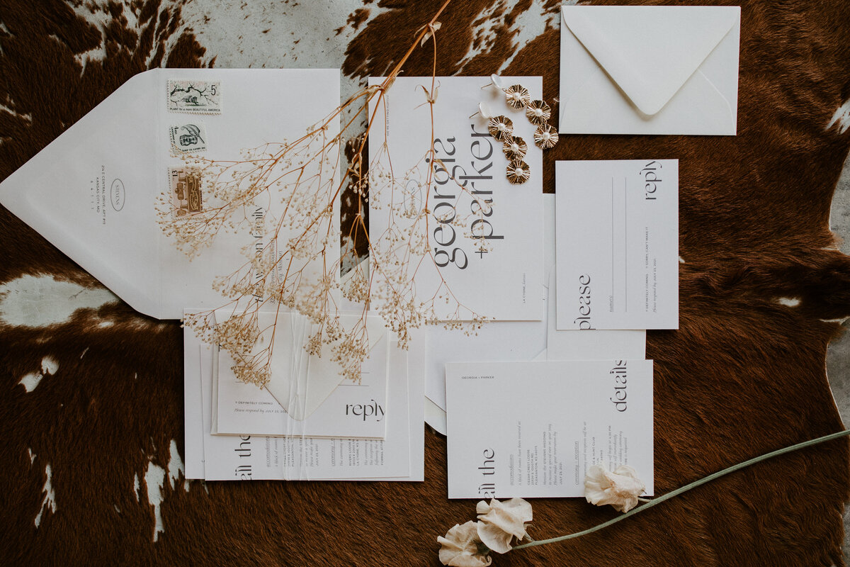 Various white wedding stationery with black font set with gold earrings and dried flower stems atop cow hide.