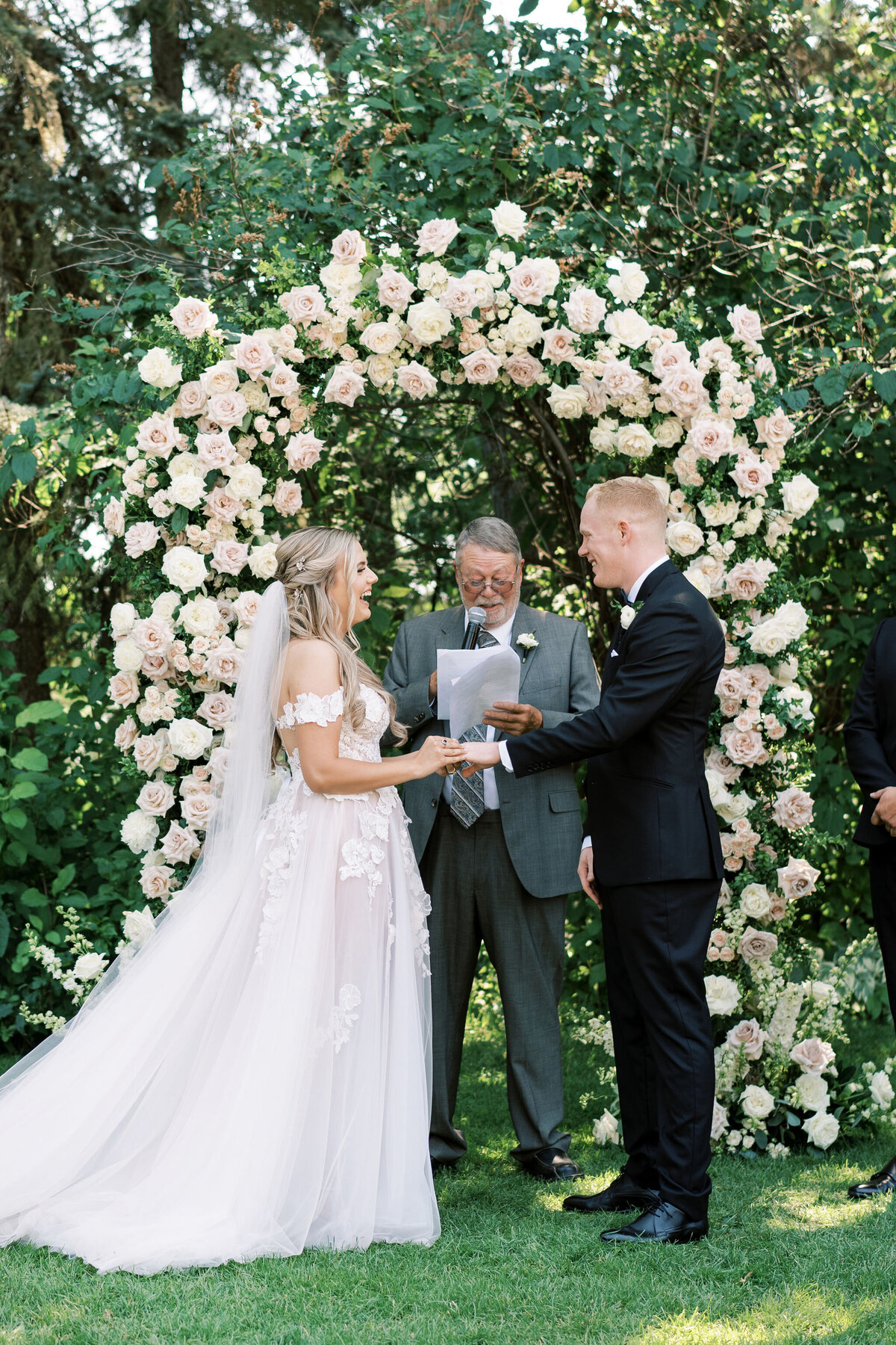 A bride in a white dress and a groom in a black suit exchange vows in an outdoor garden ceremony, standing in front of a floral arch with a person officiating. This enchanting scene unfolds at their destination wedding in Banff, Alberta.