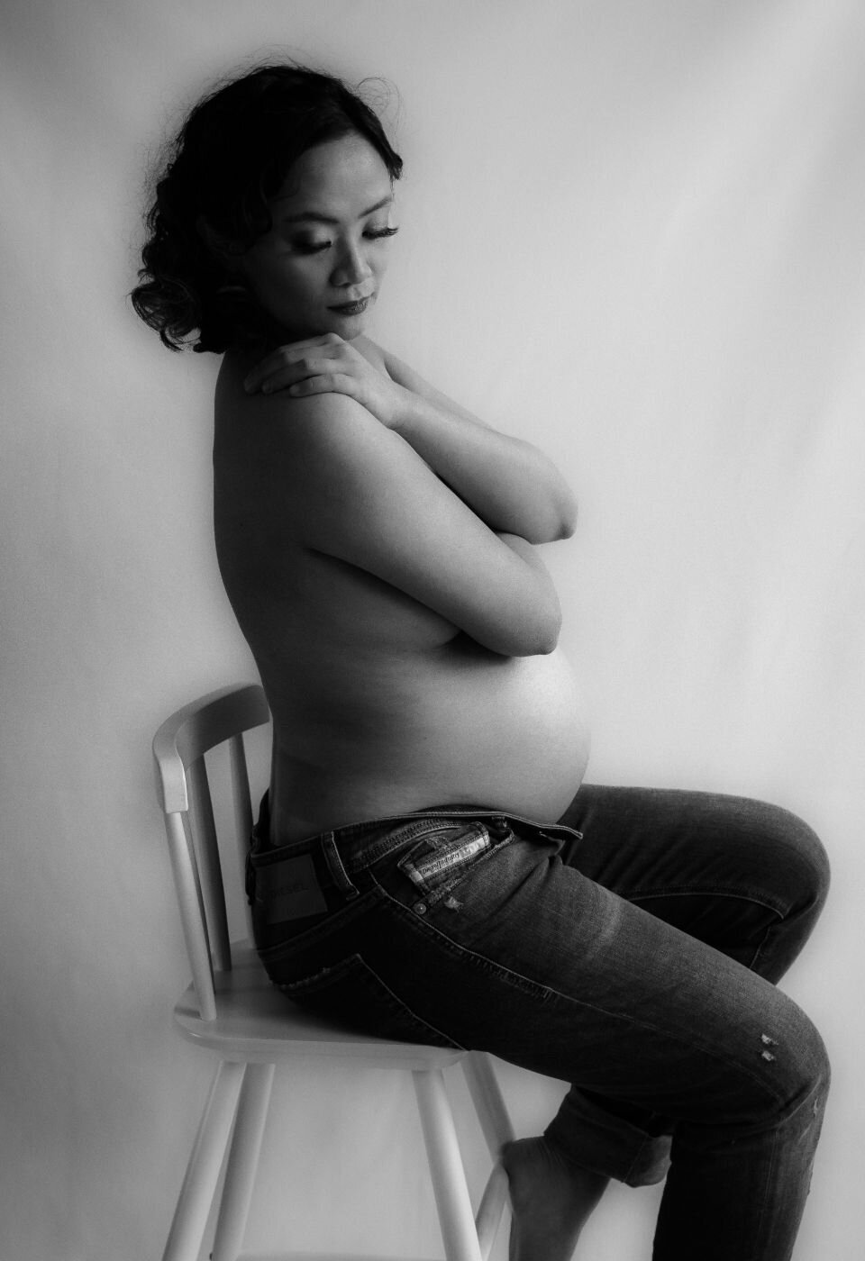 Artistic black-and-white portrait of a topless pregnant woman sitting on a chair in jeans, arms crossed over her chest in a serene pose.
