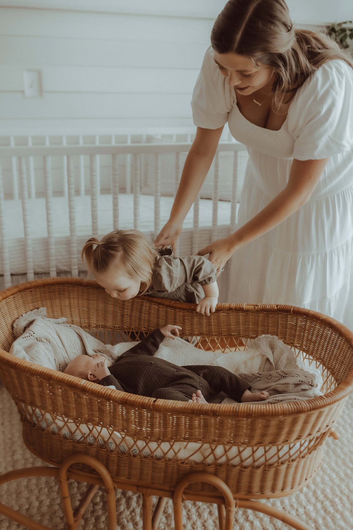 A woman in a white dress helps a toddler look into a wicker bassinet containing a baby, in a room with a crib in the background.
