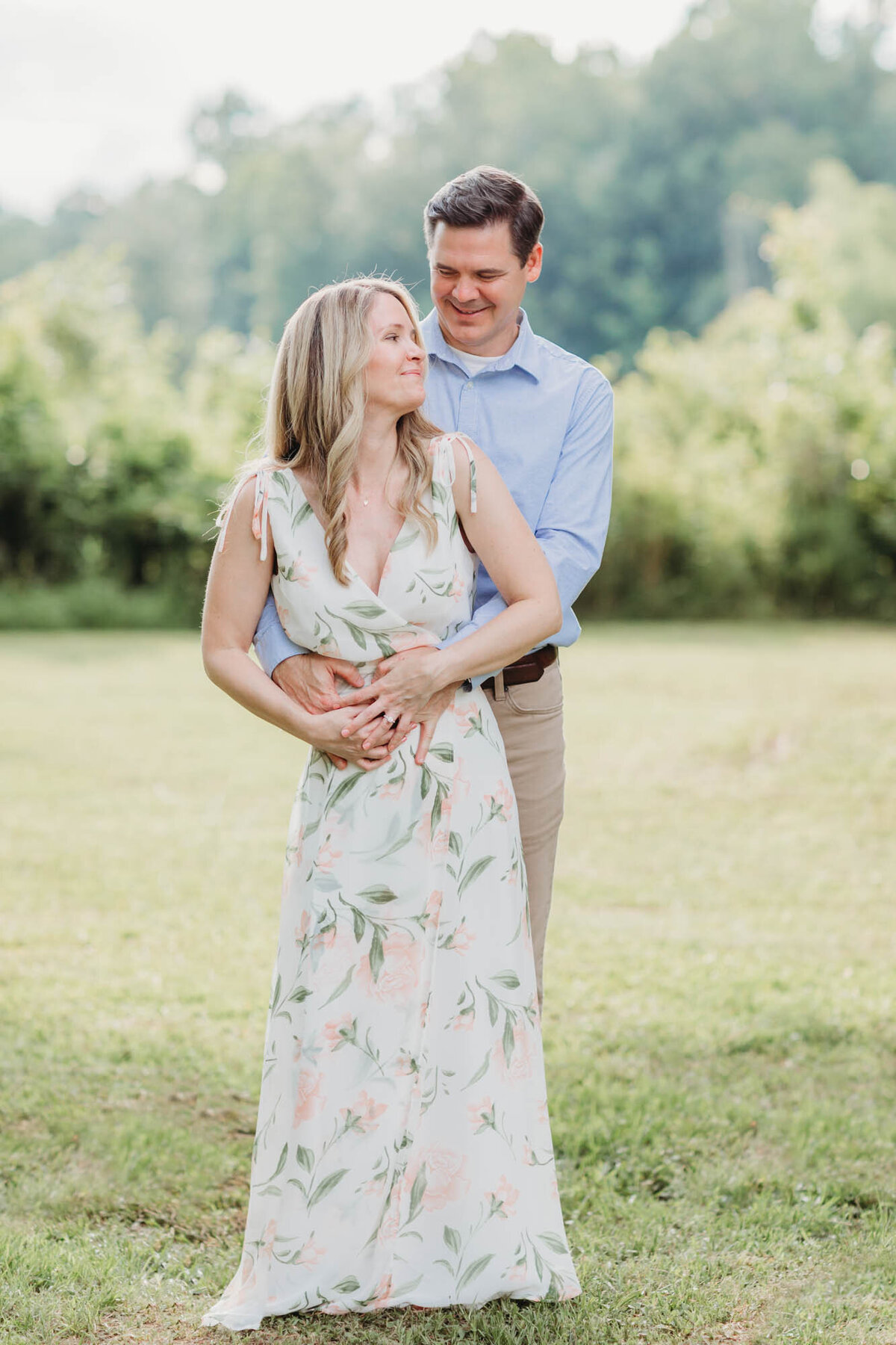 Engagement portrait of a couple on the lawn with her standing in front of him and he is looking at her.
