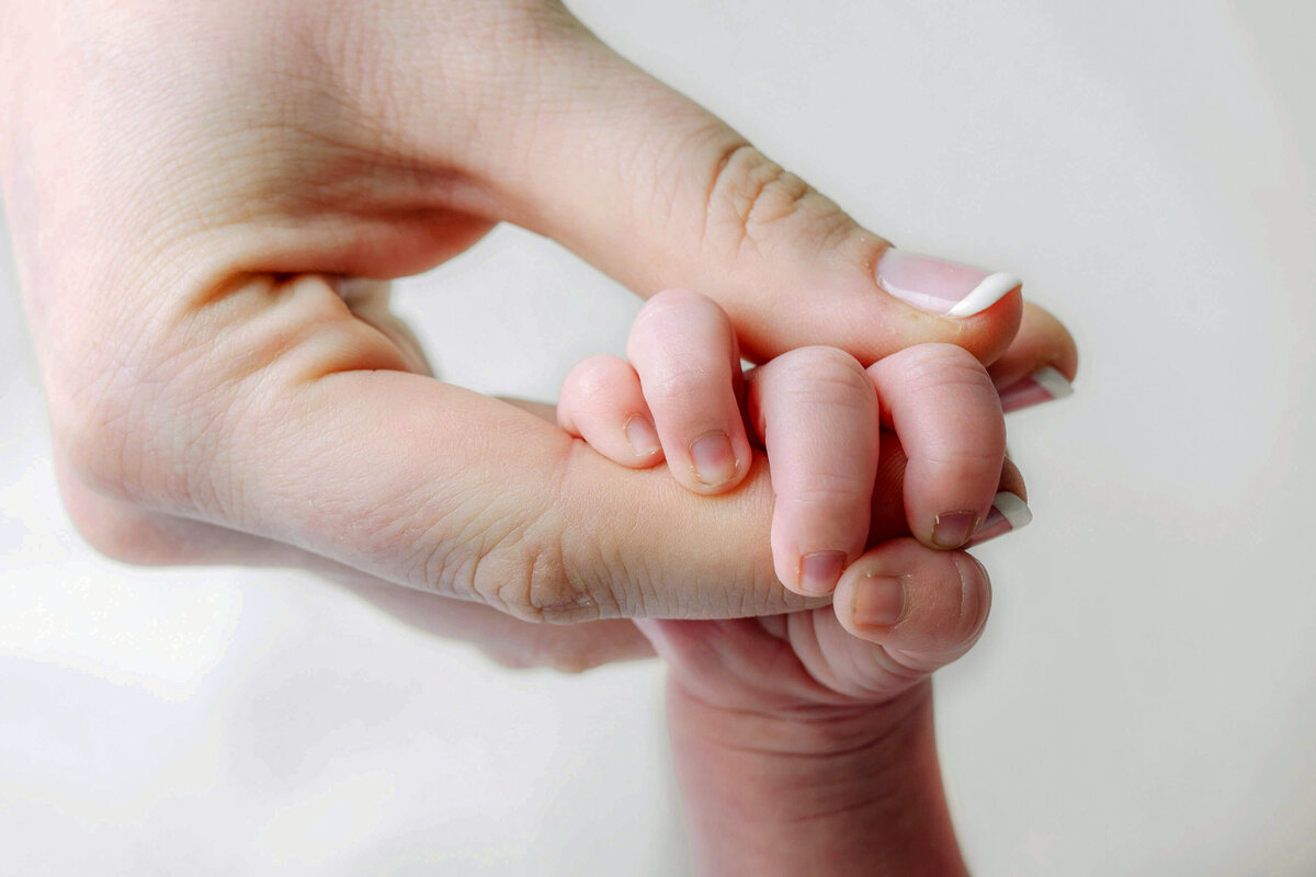 A mom is holding her newborn baby's hand while the baby is gripping his mom's finger on a white backdrop.