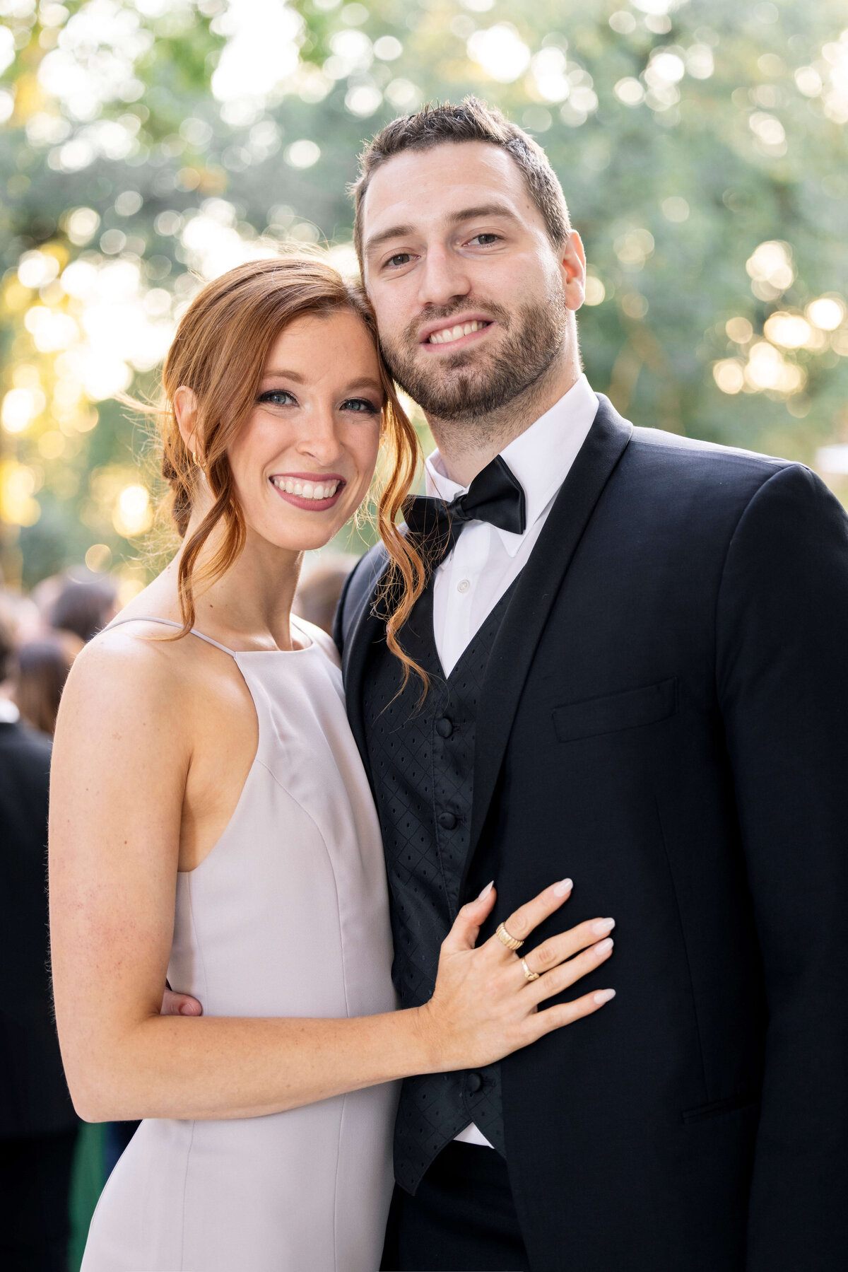 A couple dressed formally, standing close together. The woman is wearing a sleeveless, light-colored dress, while the man is in a black tuxedo with a bow tie. They are smiling at the camera, with a blurred outdoor background visible.