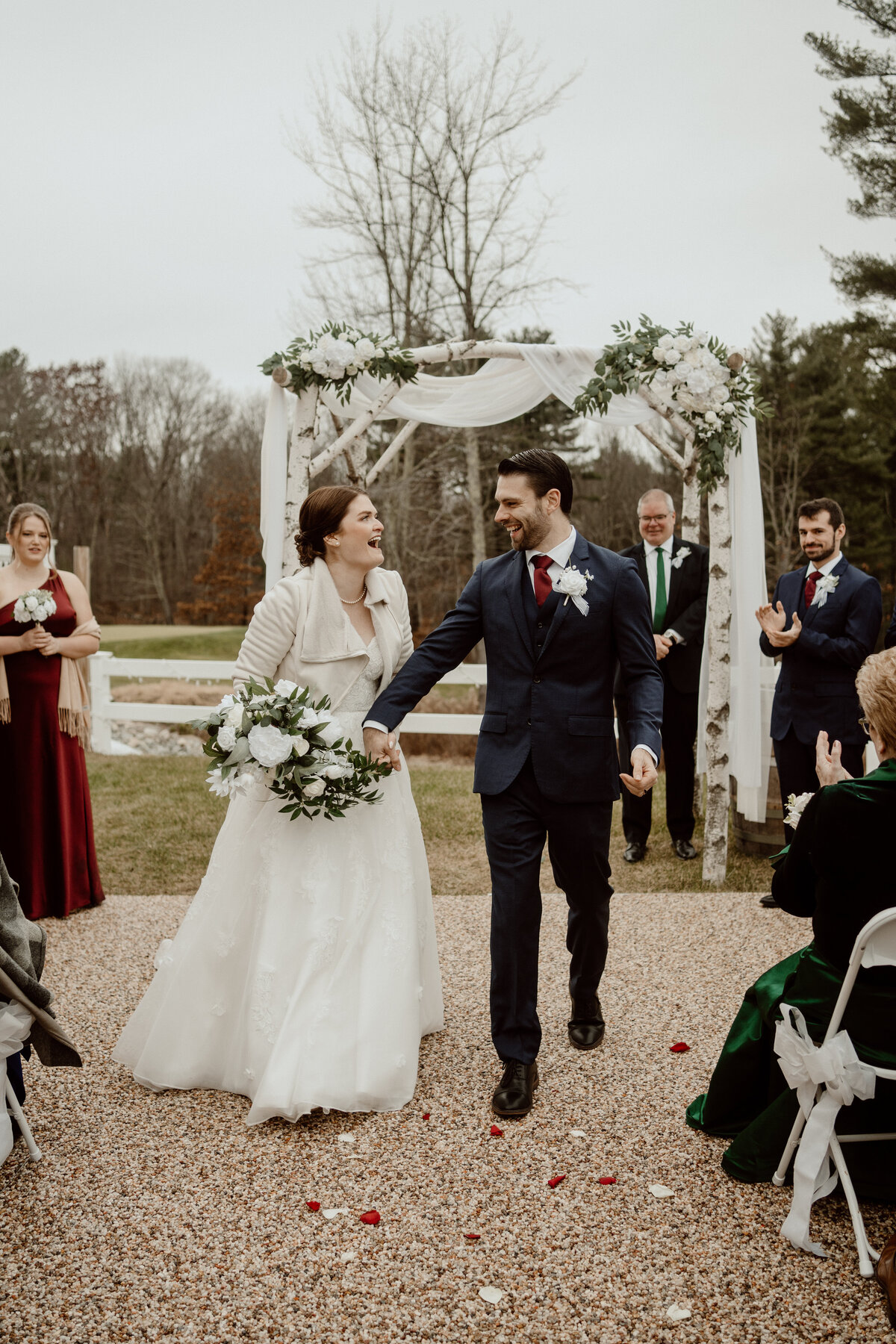 Newlywed couple joyfully walks down the aisle after their winter outdoor wedding ceremony at Barn at Blackstone Nationalunder a white floral arch. The bride wears a white gown and a cozy jacket, holding a bouquet of white flowers, while the groom looks dapper in a navy suit and red tie. Guests and wedding party members celebrate in the background.