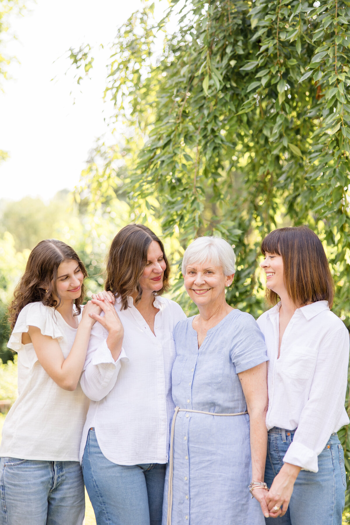 Grandma smiling at camera while daughters and granddaughter smiles at her. Wearing shades of white and blue for backyard portaits in the summer in Beavercreek, Ohio.