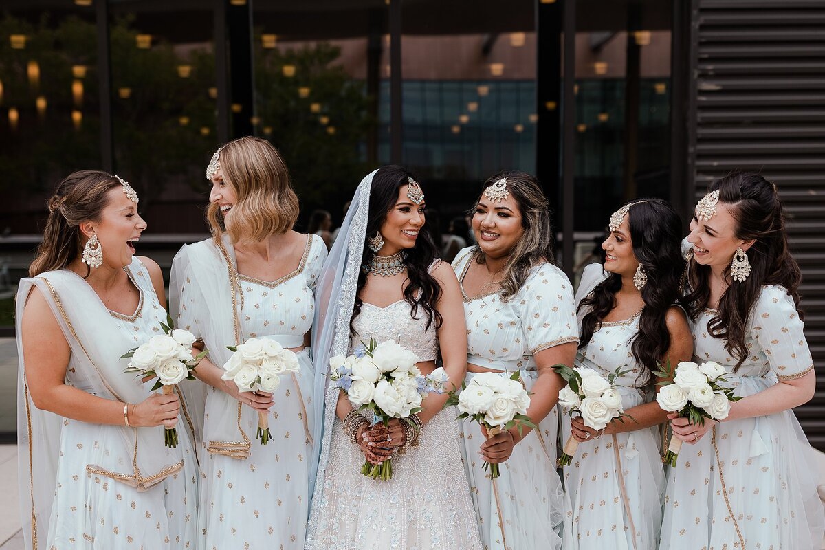 The Indian bridesmaids wearing light blue and gold sarees are holding small white bouquets as they laugh with the Indian bride wearing a white beaded saree with a light blue and gold dupatta for her Hindu wedding in Nashville, TN