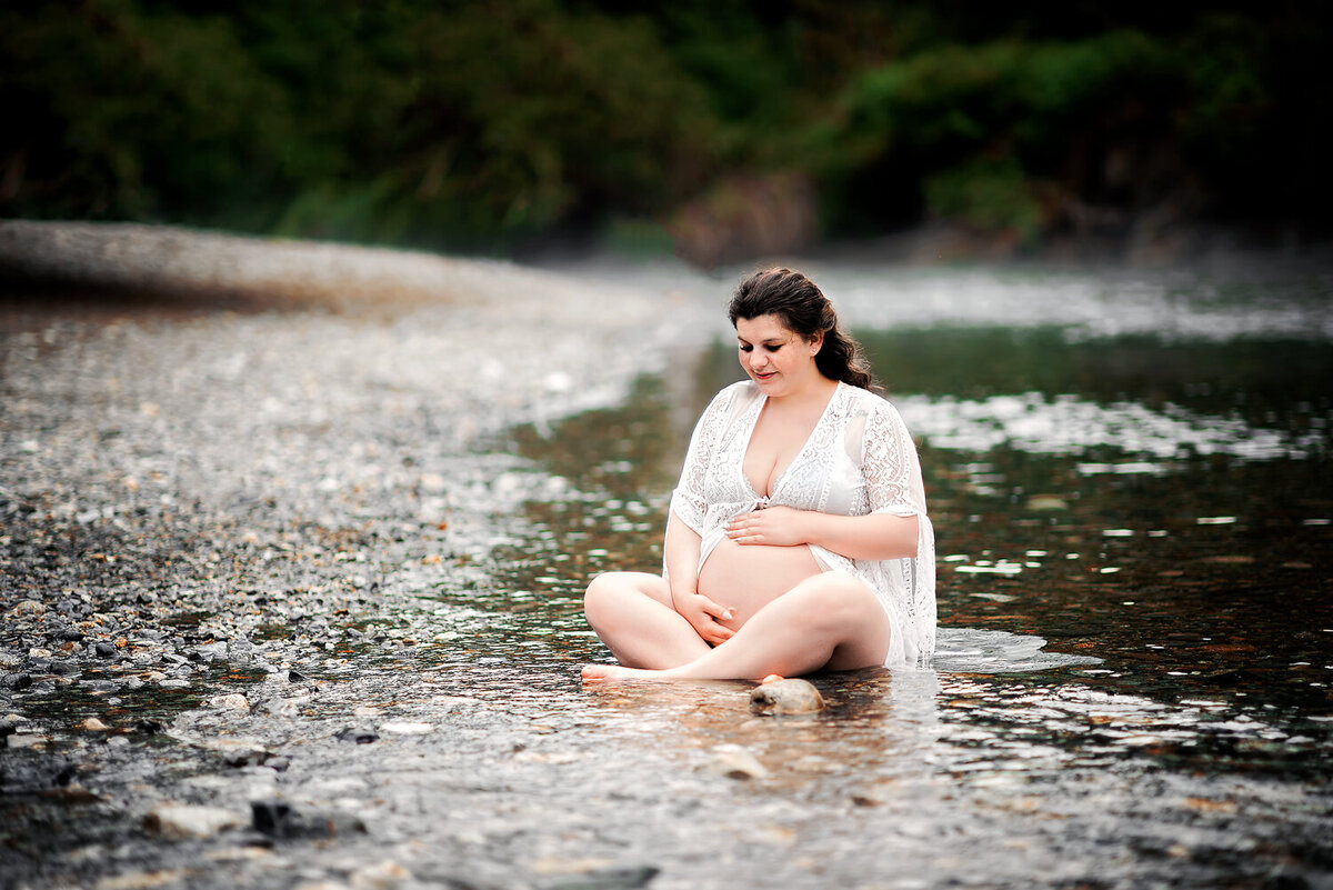 Expectant mother in white lace robe sitting in the river rocks at Golden Ears Park.
