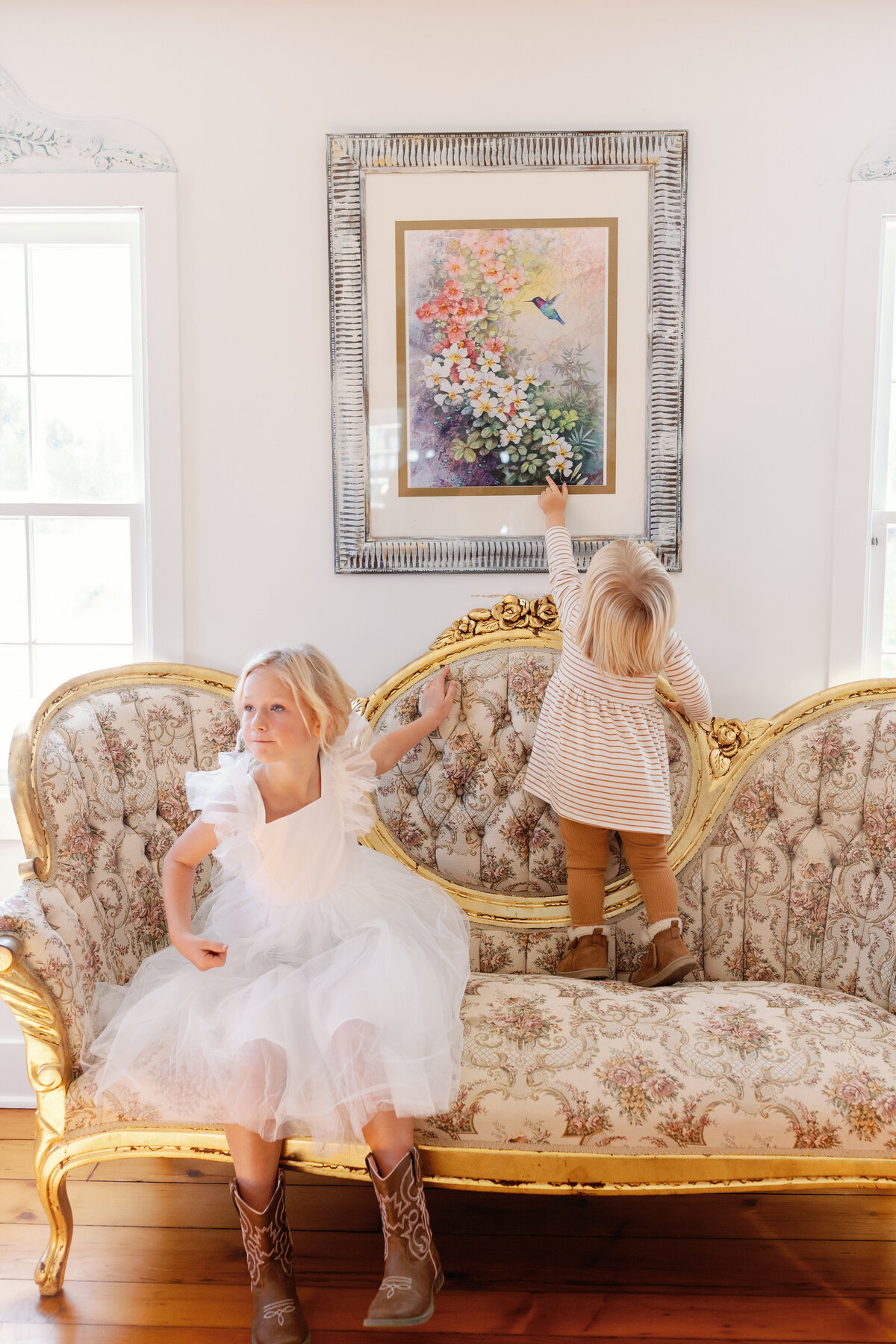 Young flower girl in white tulle dress and cowboy boots sitting on a vintage gold sofa before Maryland barn wedding.