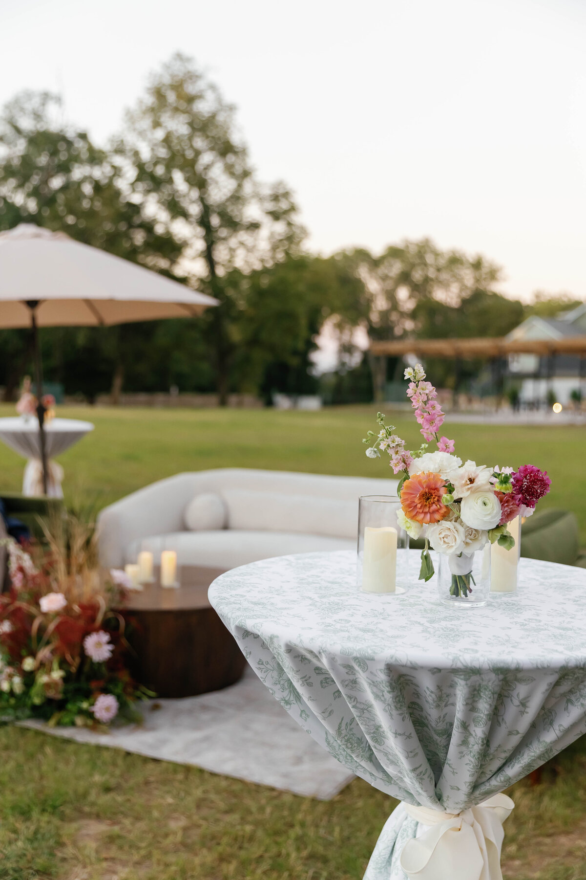 High-top cocktail table at an outdoor Arkansas wedding reception, decorated with a soft sage toile tablecloth tied with an ivory ribbon, a floral arrangement of peach, pink, and white blooms, and cylindrical candles. The setup sits in a wide grassy field at sunset with lounge seating and trees in the background, creating an elegant and romantic garden-party atmosphere.