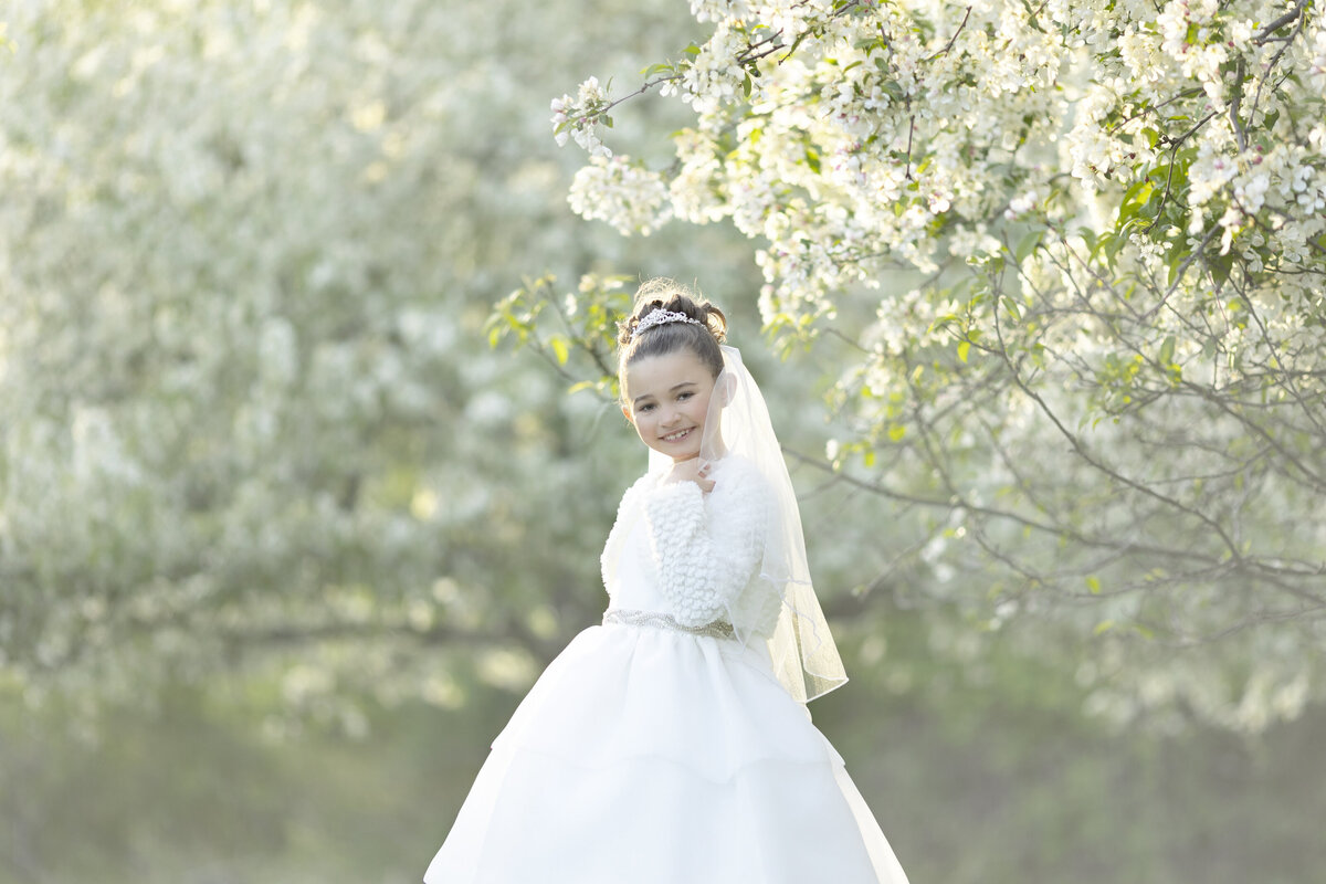 A young girl smiles under a white flowering tree at sunset in a park in a long white dress