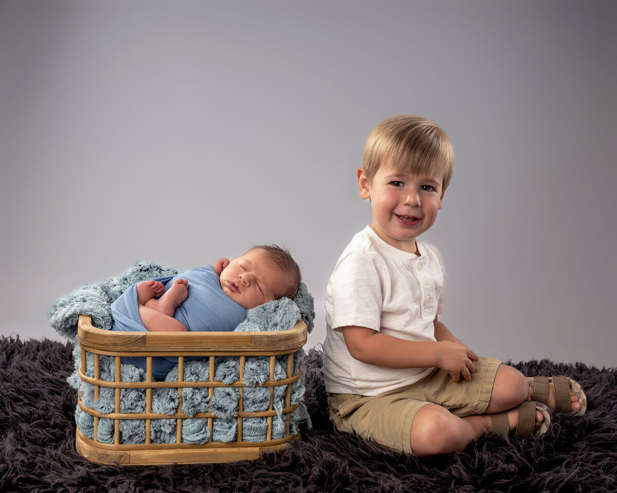 An image from an Iowa Newborn Photographer of a toddler boy in white sitting on the floor with his newborn sibling sleeping in a basket next to him