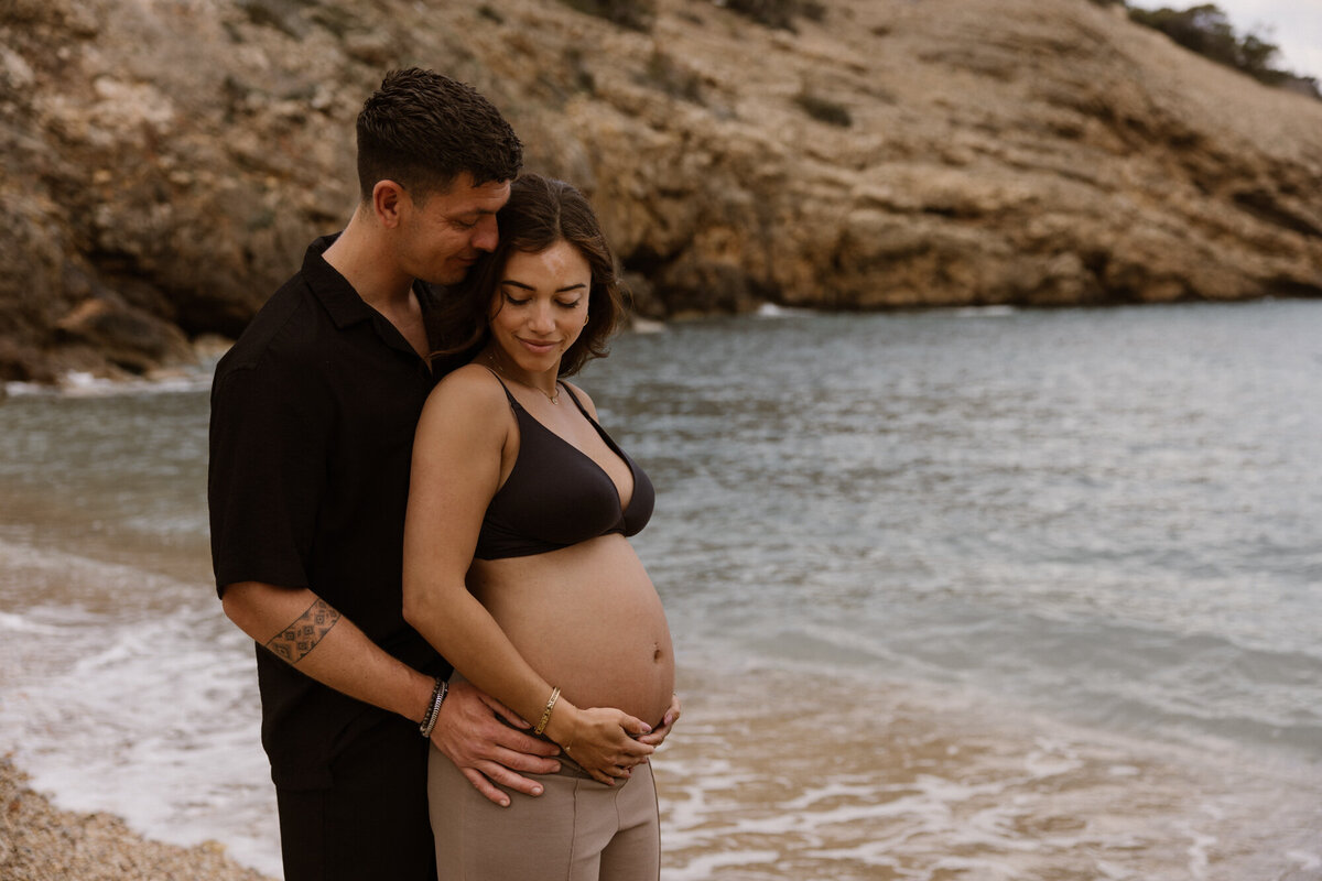 Man is standing behind a pregnant woman and hold her on a beach on Ibiza during a pregnancy shoot