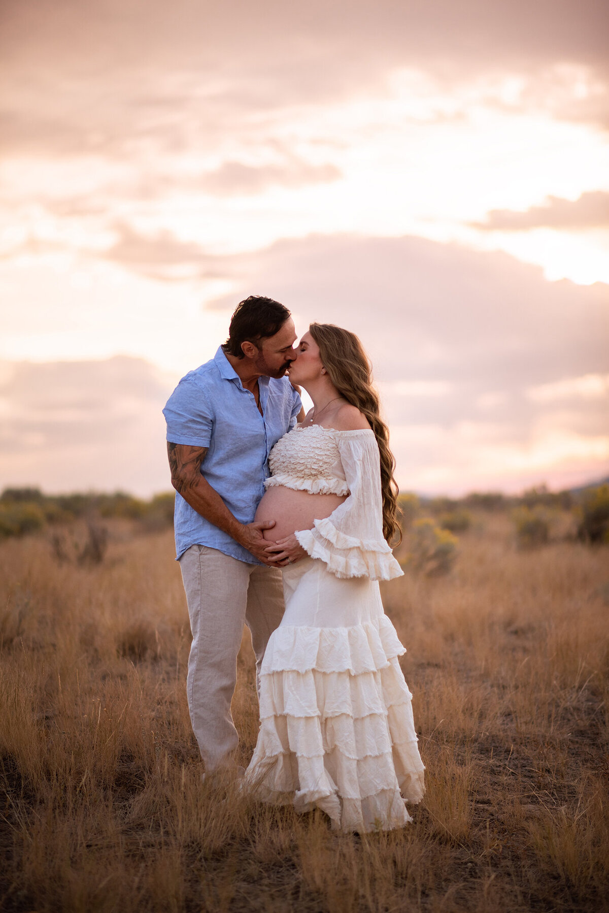 A pregnant woman wearing a mathcing cream top and skirt with her belly exposed stands next to her husband who is caressing her belly. Taken by Brittany Danielle with a beautiful sunset in the background.