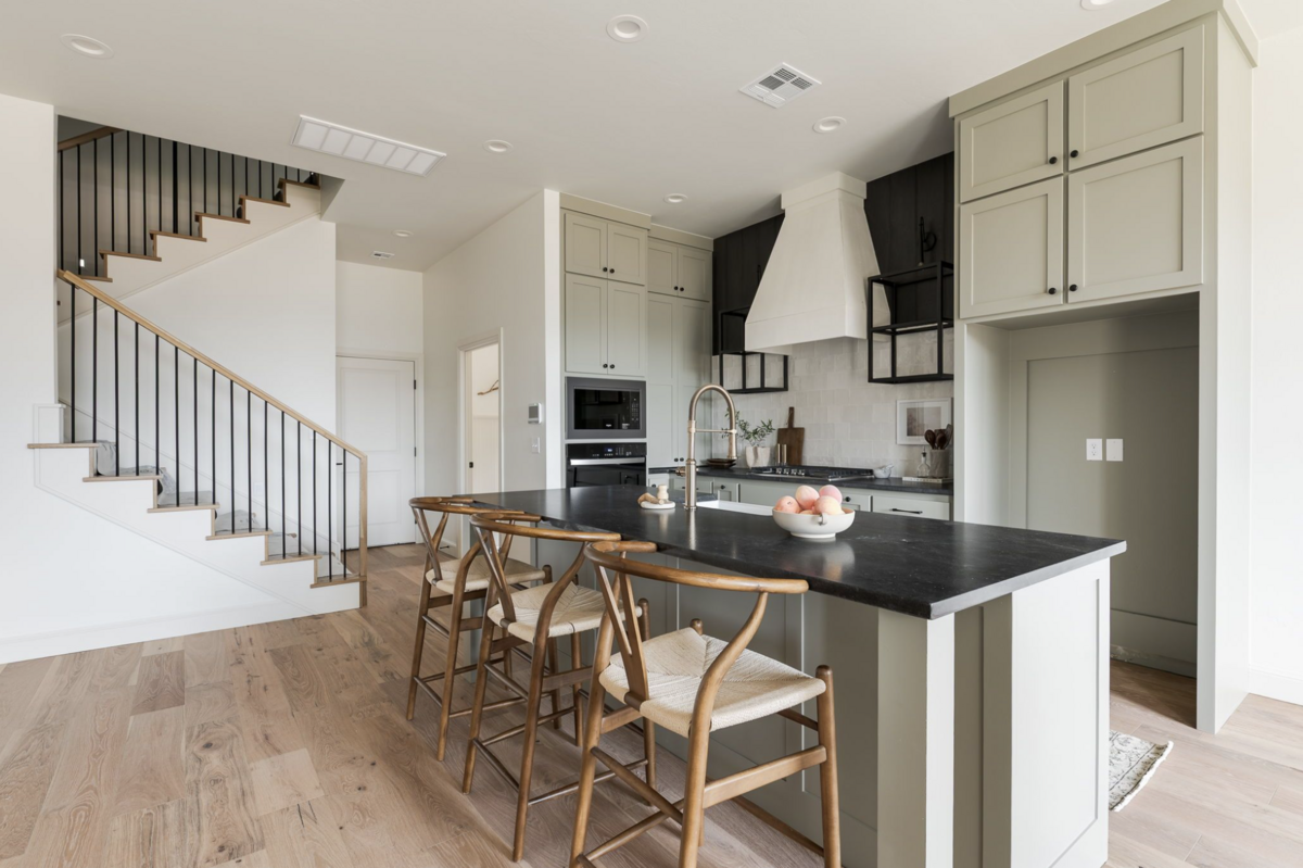 Light-filled kitchen with sage green cabinetry, black stone countertops, wood barstools, and a custom plaster hood framed by black open shelving. Natural wood floors and a modern stair railing complete the space.