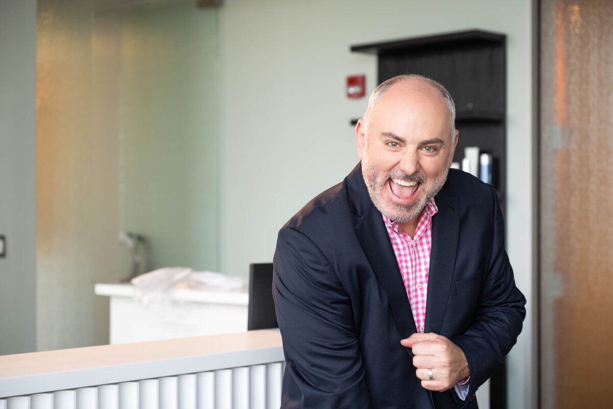 Professional headshot of a cheerful man in a navy blazer and pink checkered shirt, standing indoors with a bright and modern office background, exuding a friendly and approachable demeanor.