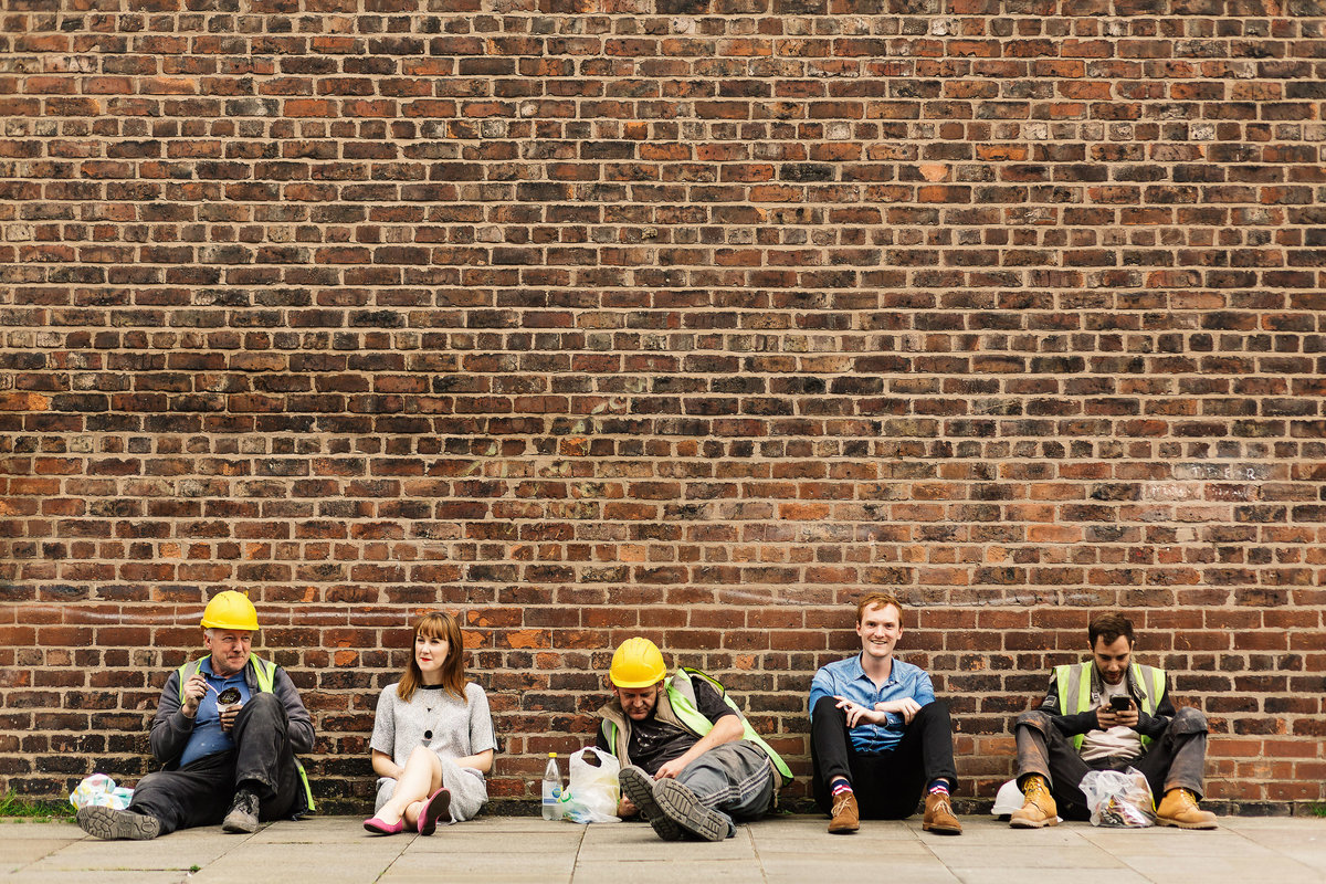 An engagement shoot photo of a couple sat against a wall in Manchester alongside some workmen having their lunch