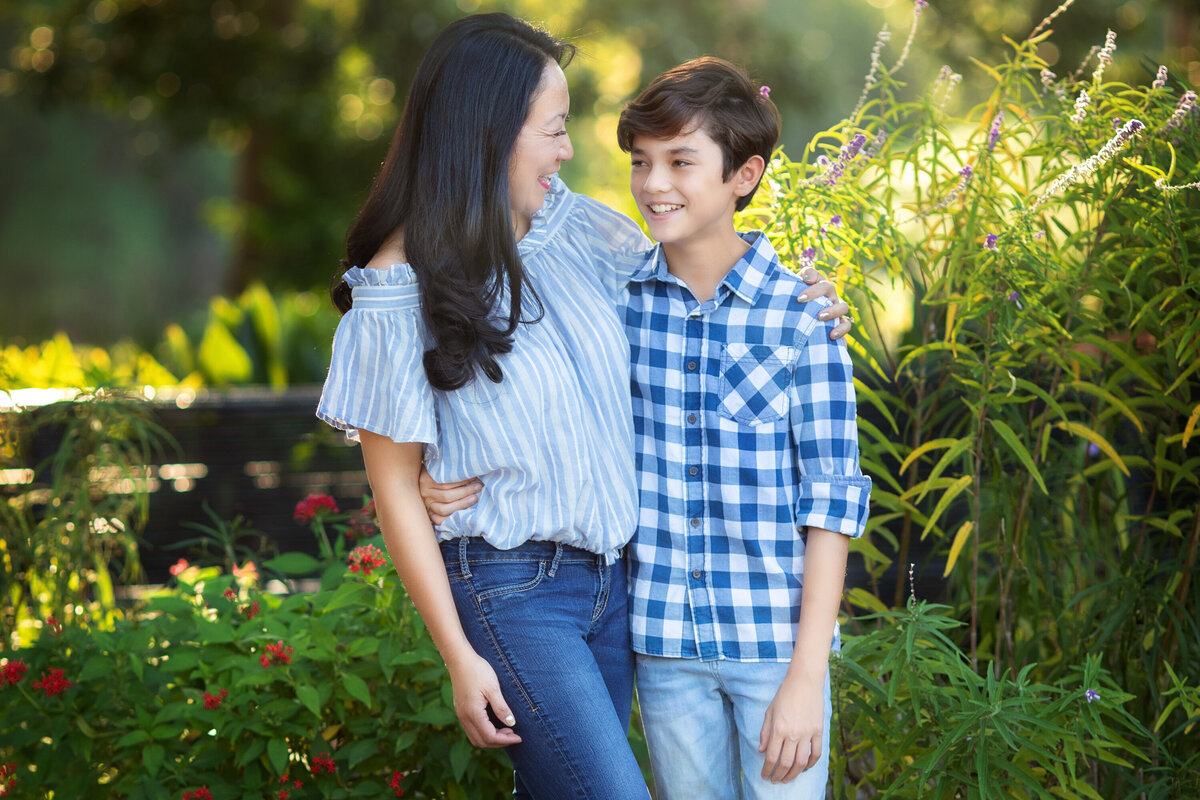 Asian mother and her 13 year old son with arms around each other.  They are wearing jeans and standing in front of flowers.  They are smiling and looking at each other.