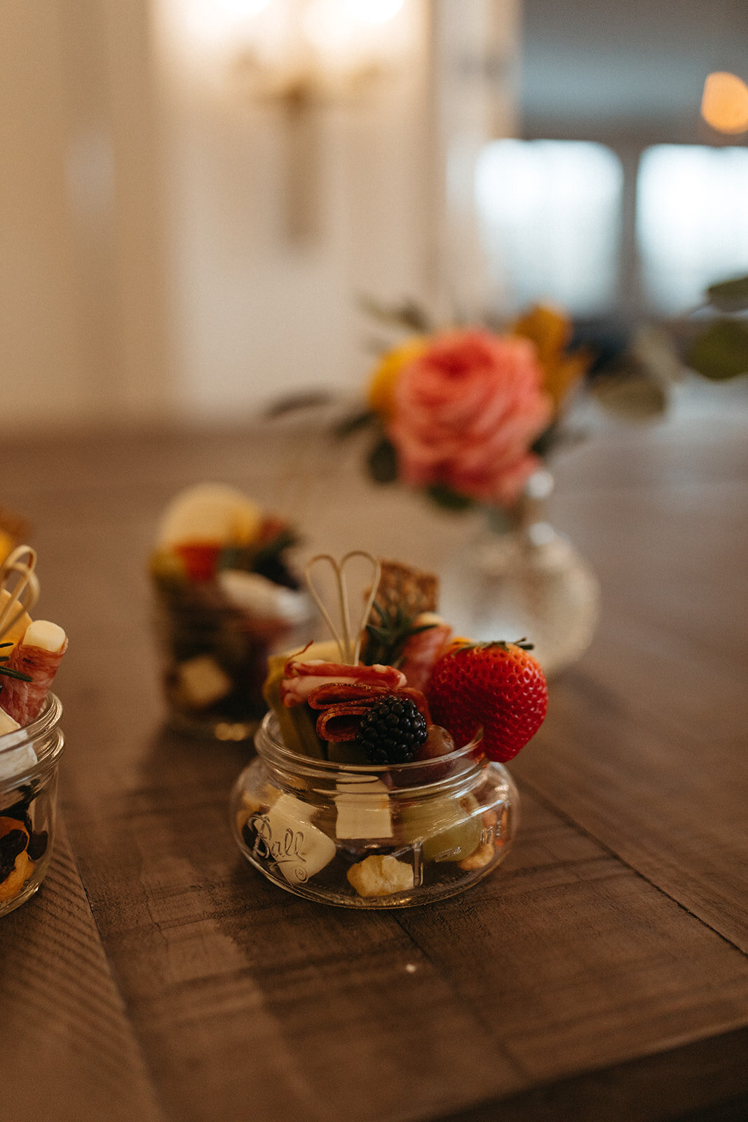 Charcuterie hors d’oeuvres in glass atop a wooden table with a rose in small vase.