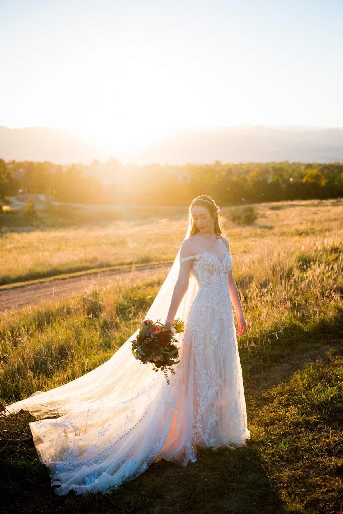 Bride stands in a field at sunset staring down over her shoulder at her bouquet.