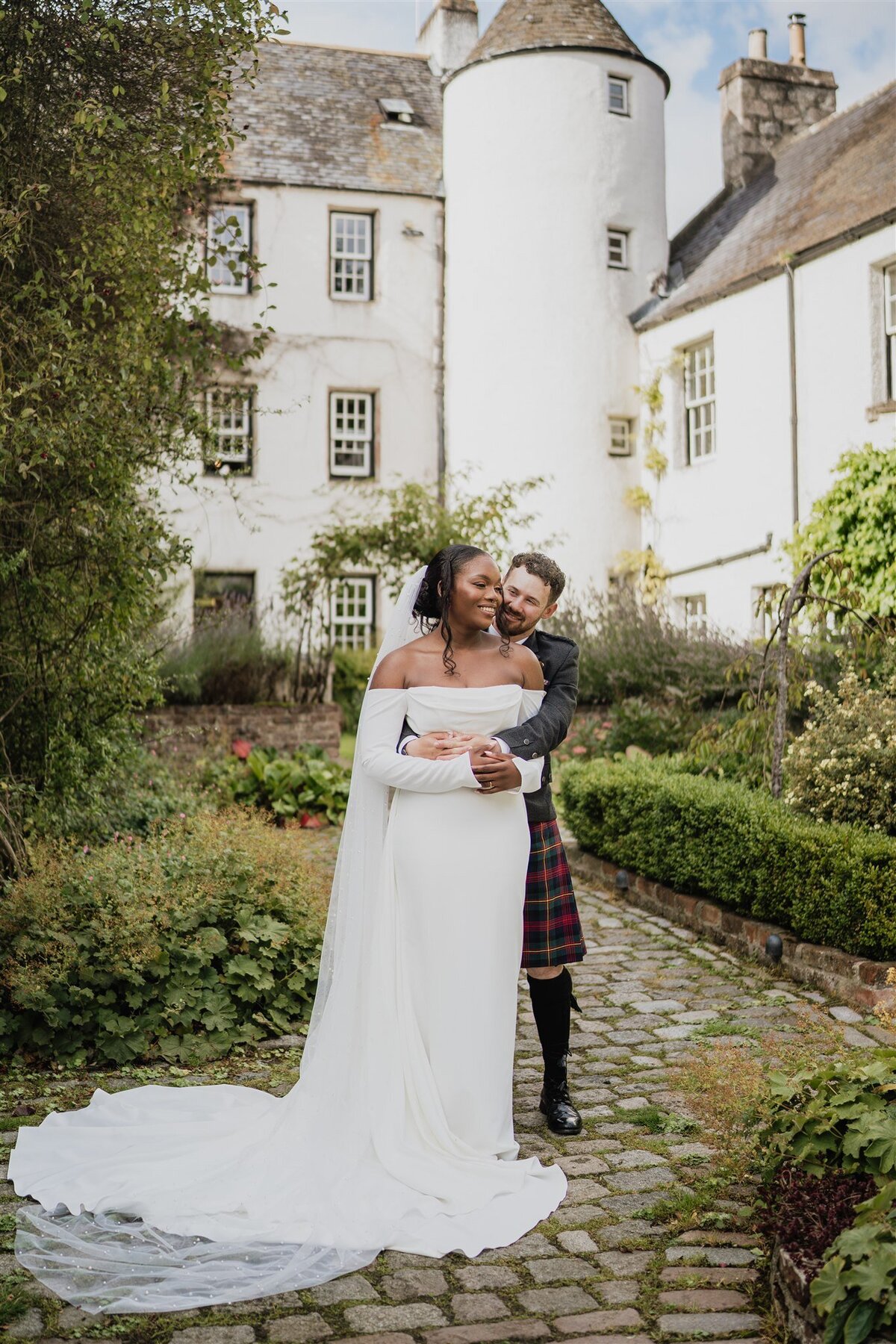 A groom wraps his arms around his wife from behind. They stand in a shaded courtyard and the white exterior of Logie Country House can be seen all around them. They stand on a cobbled surface and pose for wedding photos.