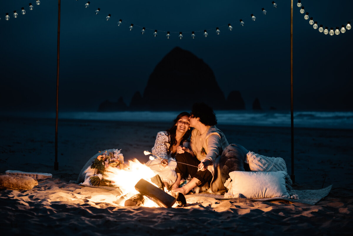 At the end of their Oregon elopement, a couple snuggles up by a bonfire on cannon beach. The shadow of Haystack rock stands in the background