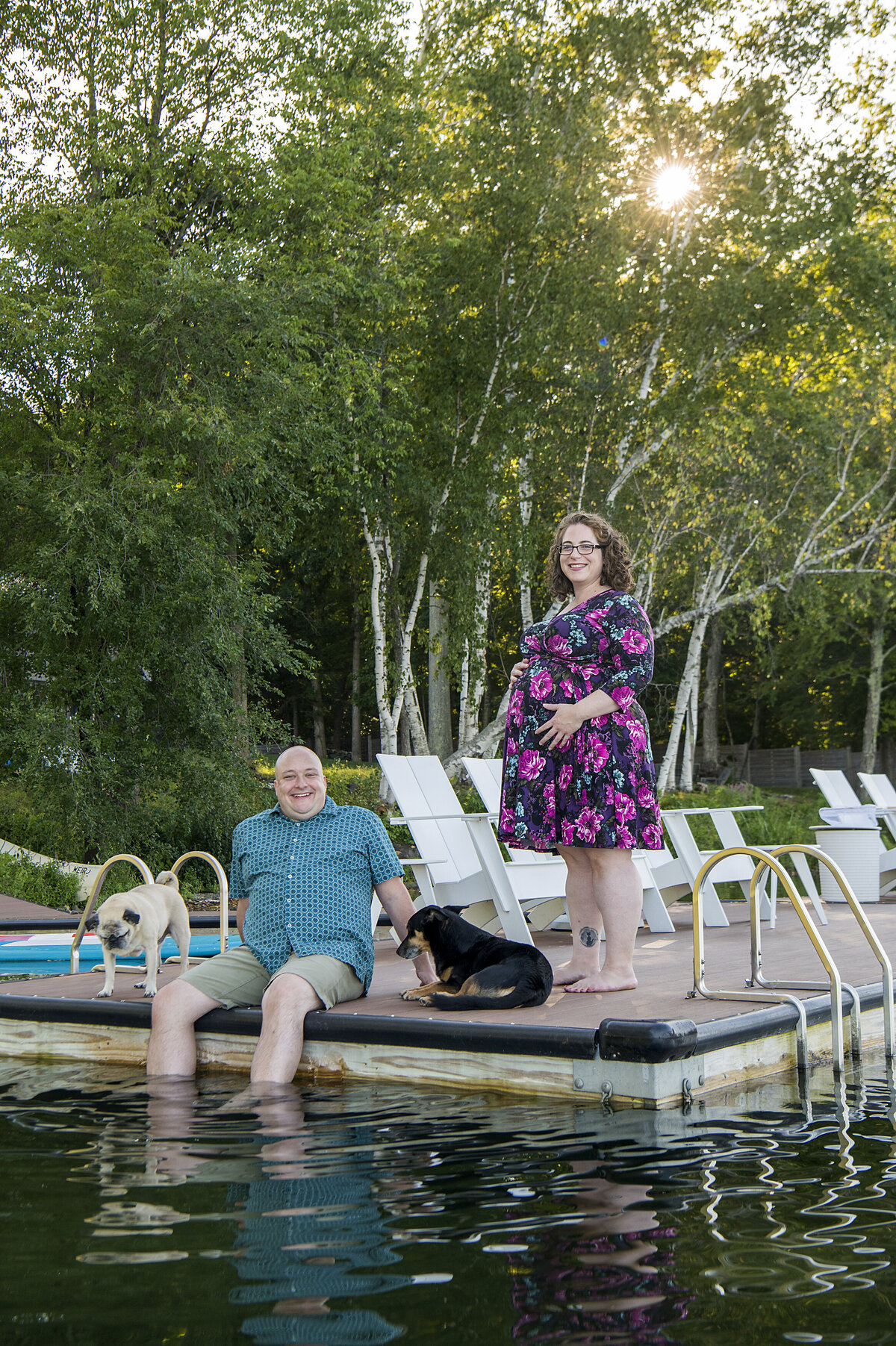A pregnant couple standing and sitting at the edge of a pool with two dogs 