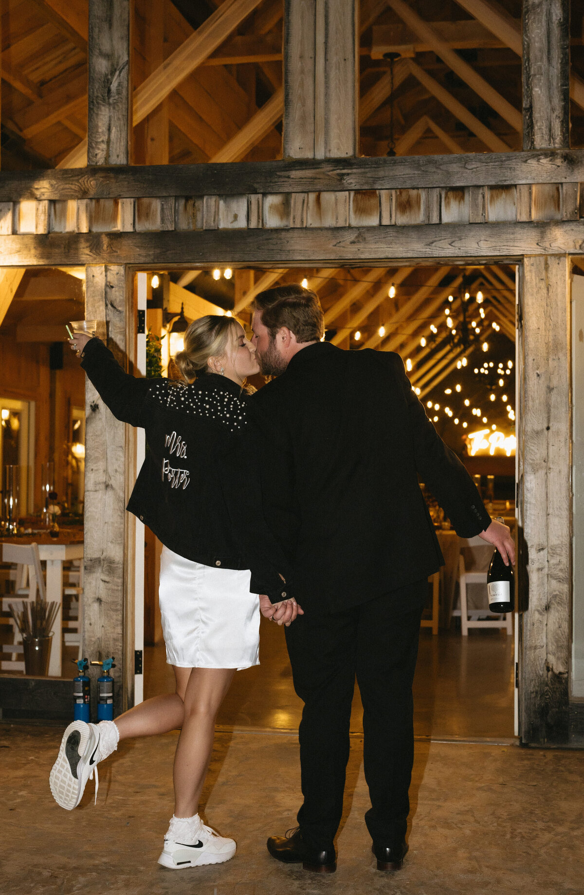 wedding reception in a barn with over head beams covered in fairy lights to create a romantic ambiance as the bride and groom hold hands and walk into their reception with their arms in the air