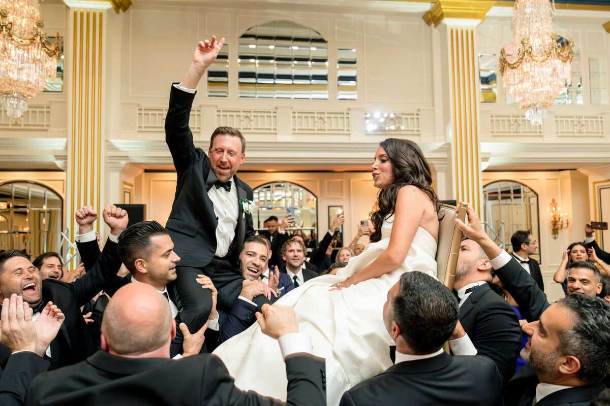 A joyous wedding celebration with a man and woman being lifted on chairs by a crowd of people in a grand ballroom. The man raises an arm in excitement. The room features large chandeliers and mirrors, and everyone is dressed in formal attire.
