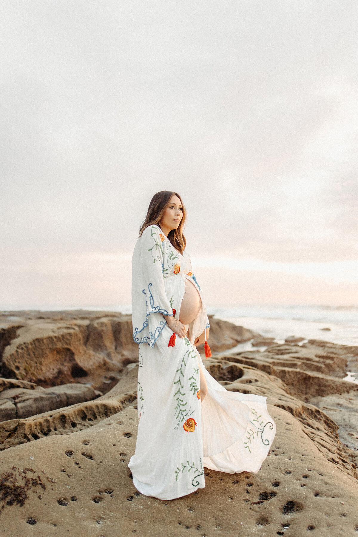 Expecting mother in a flowing white embroidered gown standing on the rocks at La Jolla during a coastal maternity session. Captured by San Diego Family Photographer Yuka Schad.