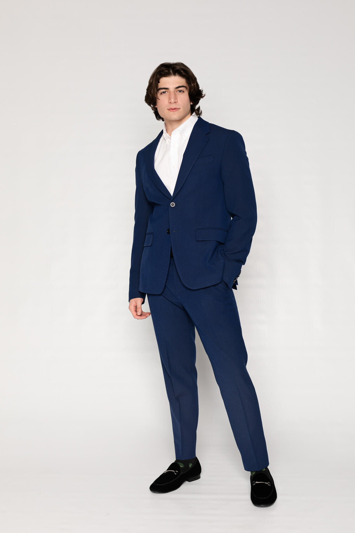 A person with medium-length hair stands confidently, wearing a dark blue suit with a white dress shirt and black shoes. The background is plain white, accentuating the formal attire.
